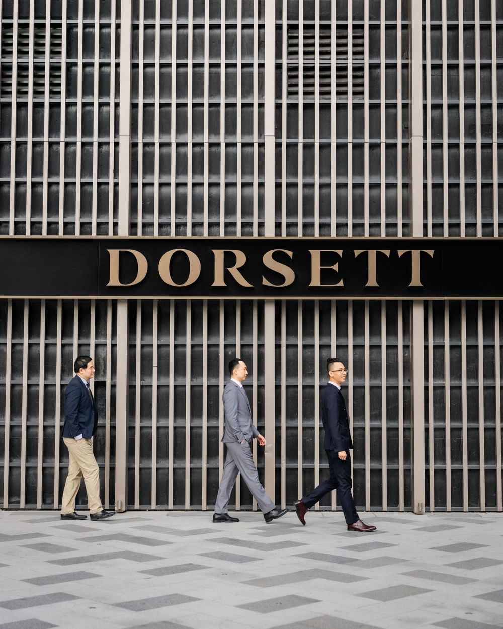 Three men in formal suits walking past the Dorsett hotel building facade.
