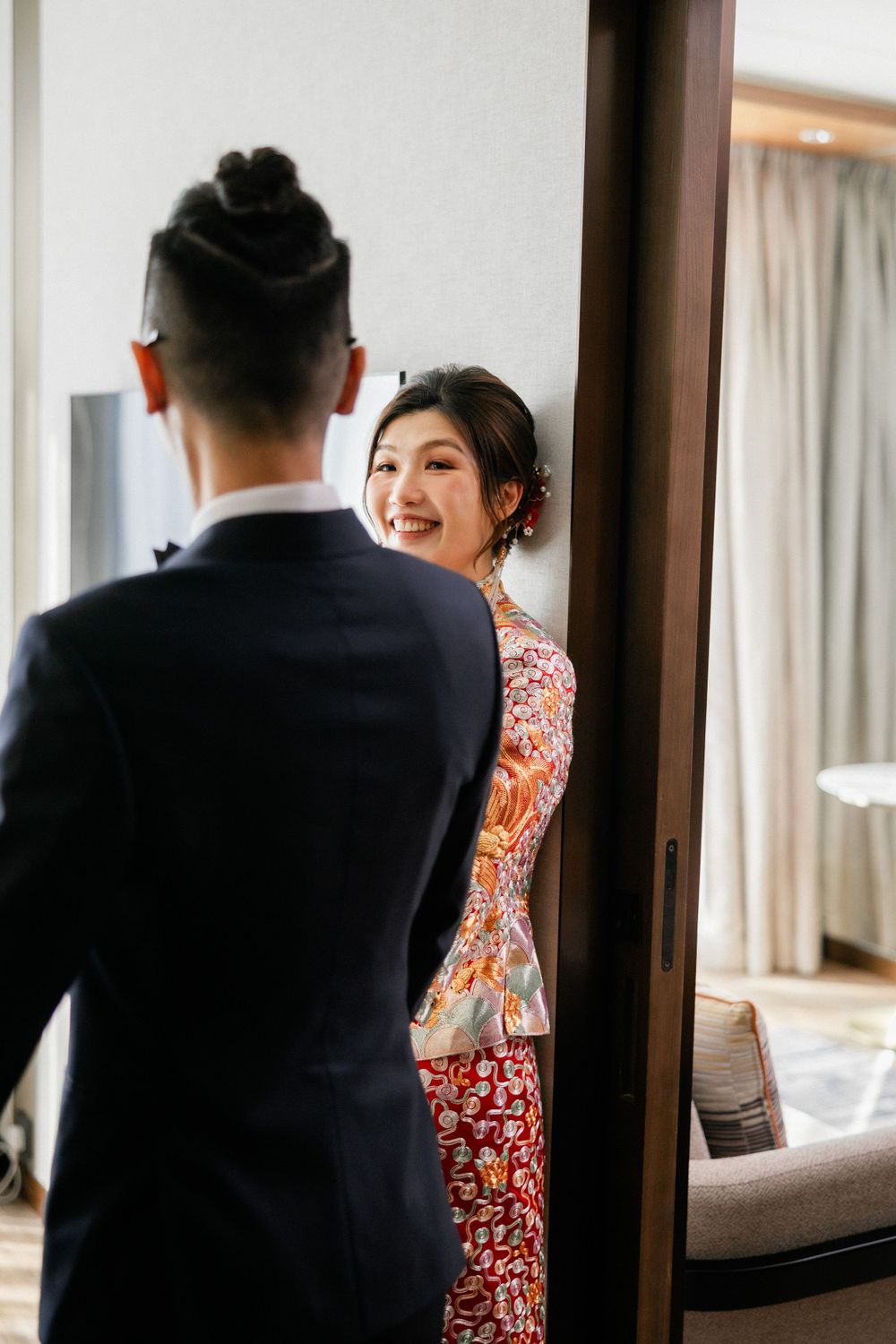 Smiling bride in red floral qipao peeking around a mirror at groom in black suit.