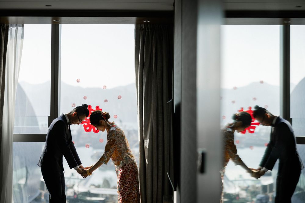 Couple in traditional Chinese wedding attire holding hands by window with city view reflection.