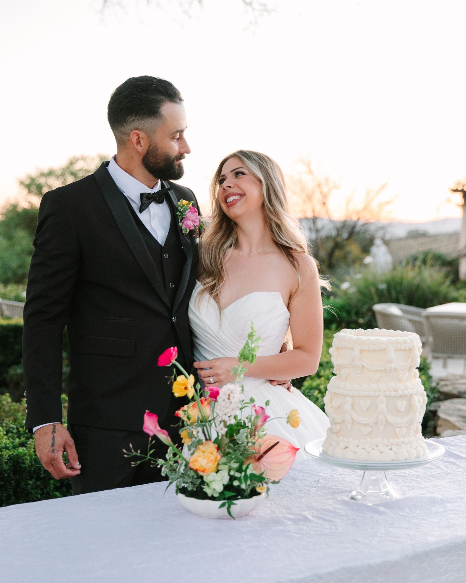 A couple shares an intimate moment while cutting their elegant white wedding cake at an outdoor reception.