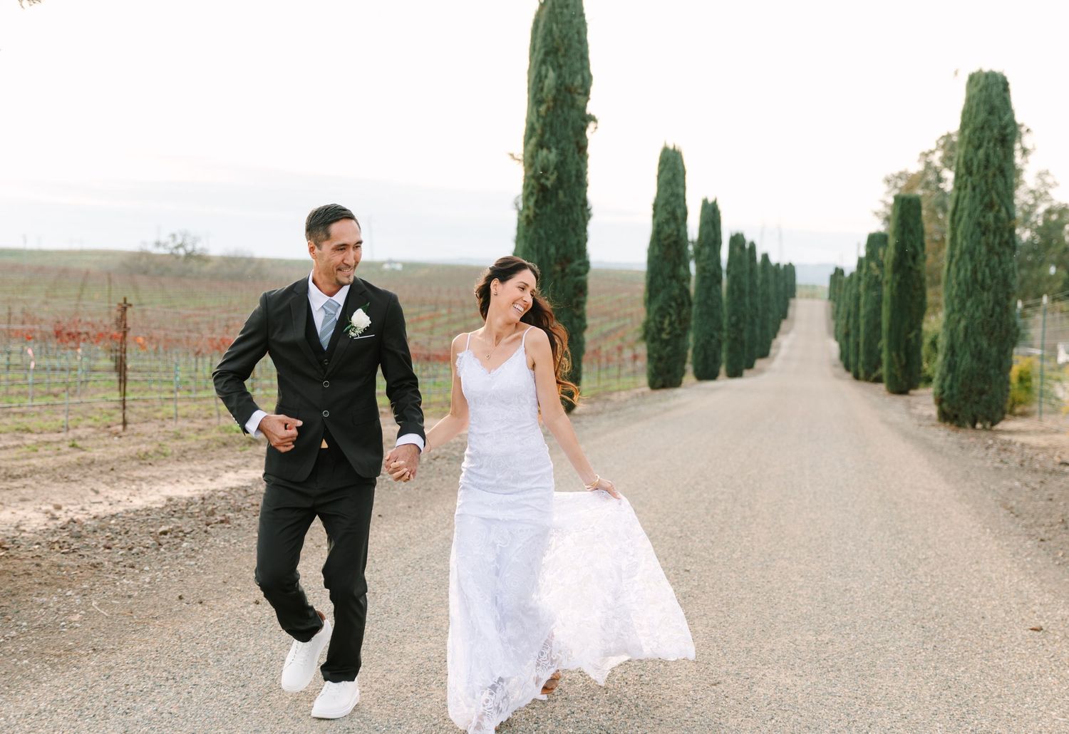 Wedding couple runs playfully down a cypress tree-lined road in the countryside.