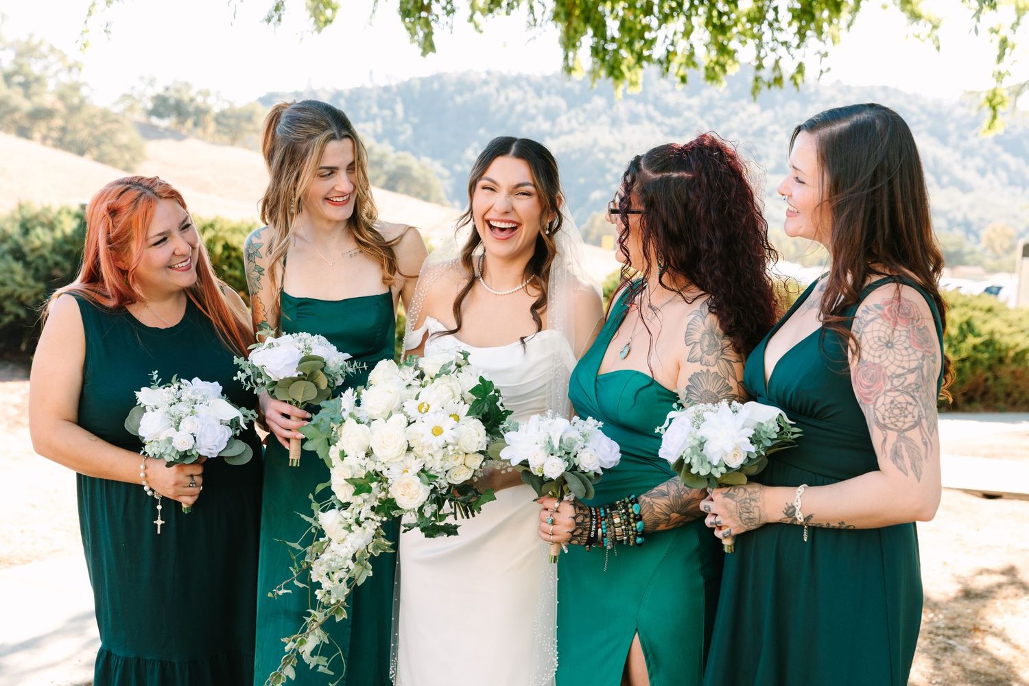 Wedding party in emerald green dresses holding white bouquets pose together outdoors on a sunny day.
