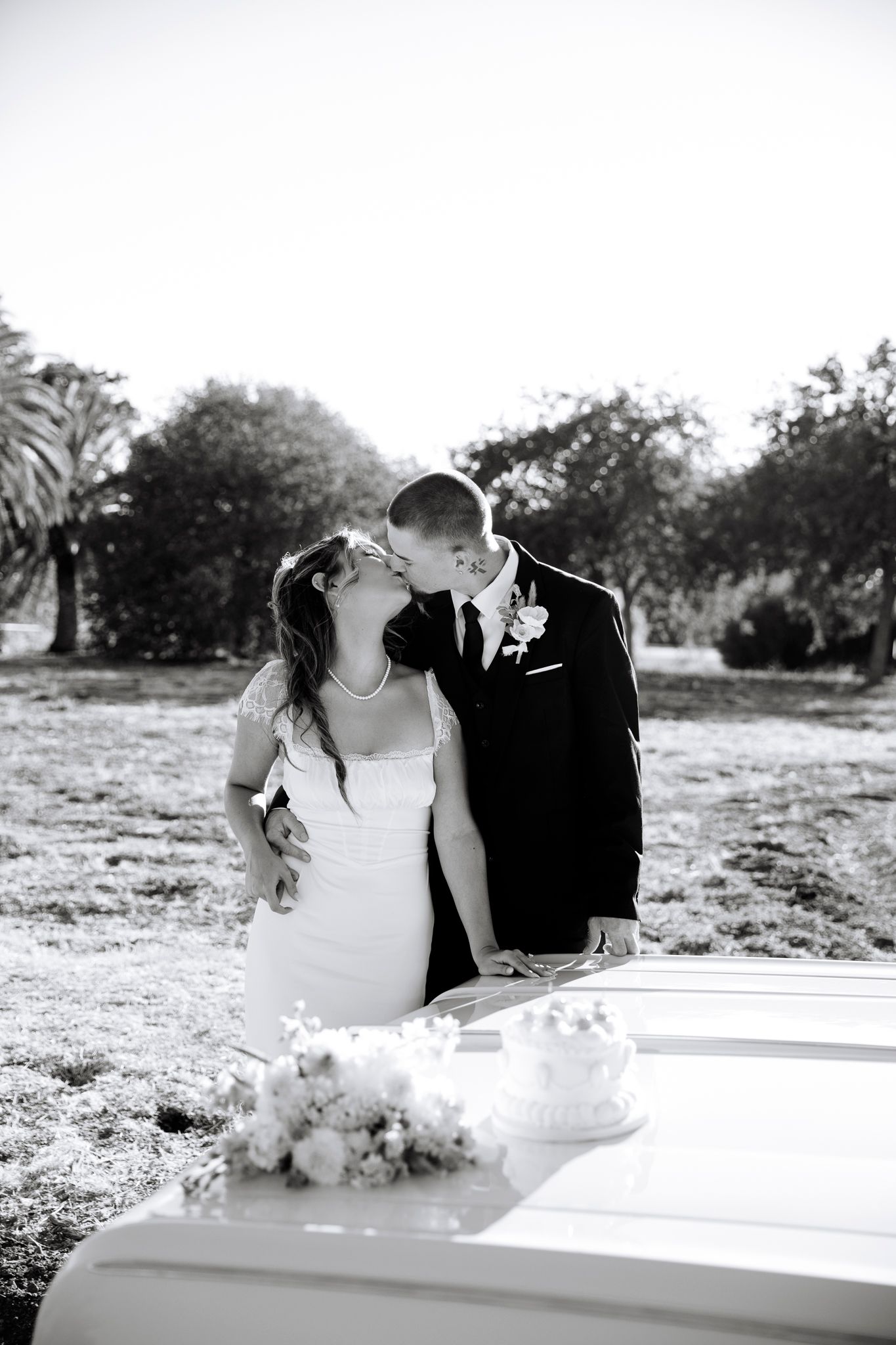 A black and white wedding photo shows a loving couple sharing a kiss next to their cake table outdoors.
