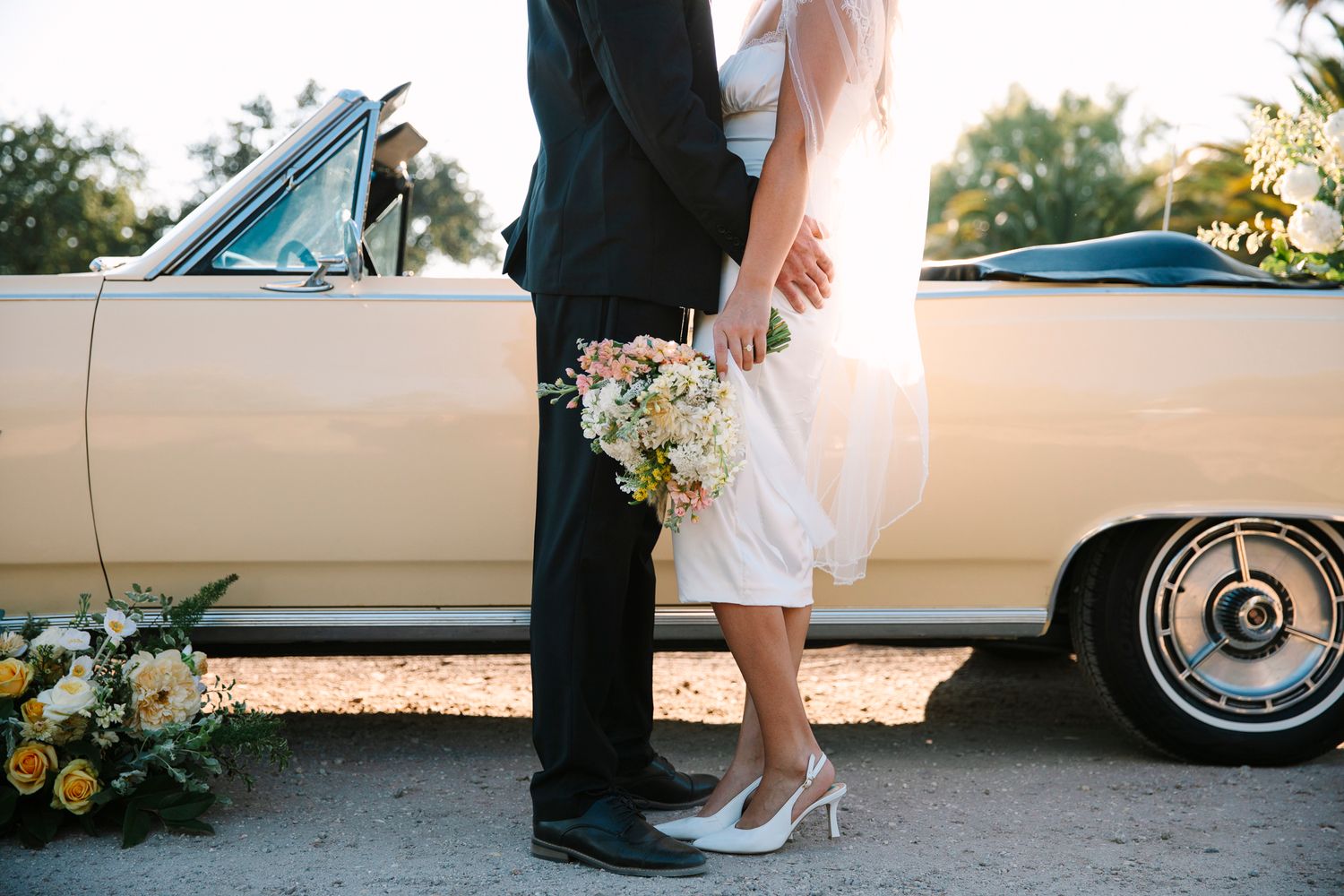 A romantic wedding photo showing elegant bridal shoes and a floral bouquet next to a vintage cream-colored car.