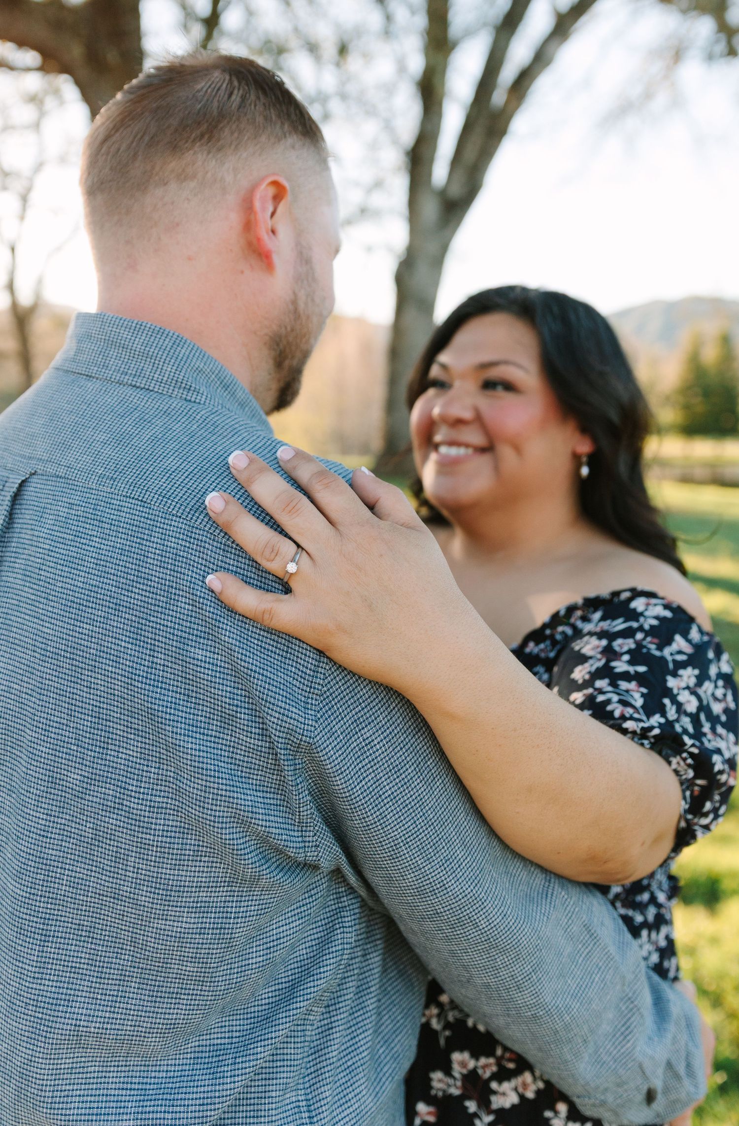 A couple shares a romantic moment outdoors in a park, with the person in a gray sweater embracing their partner in a floral dress.