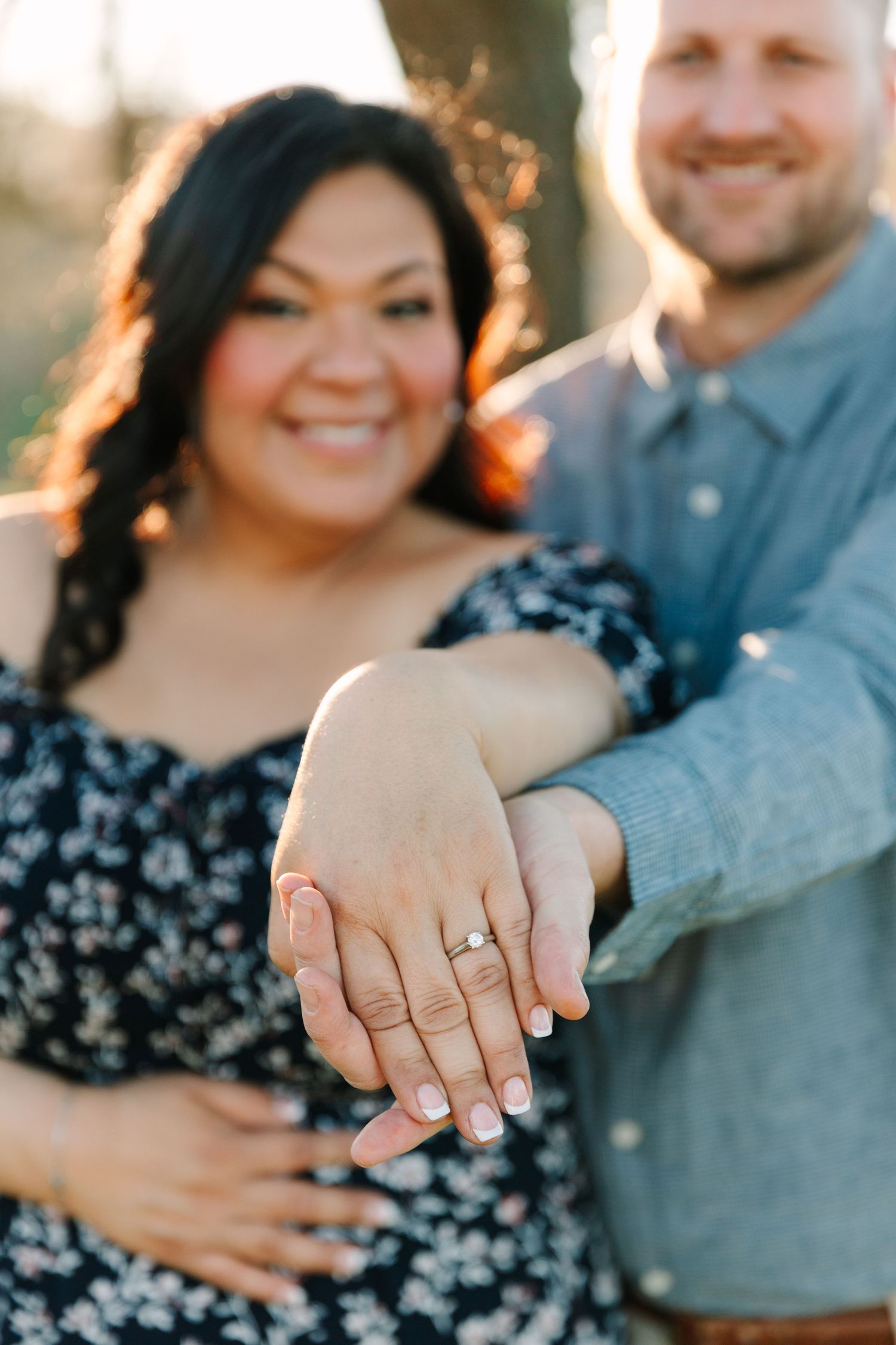 A couple shows off their engagement ring in a sunlit outdoor setting, with a floral dress and denim shirt visible.