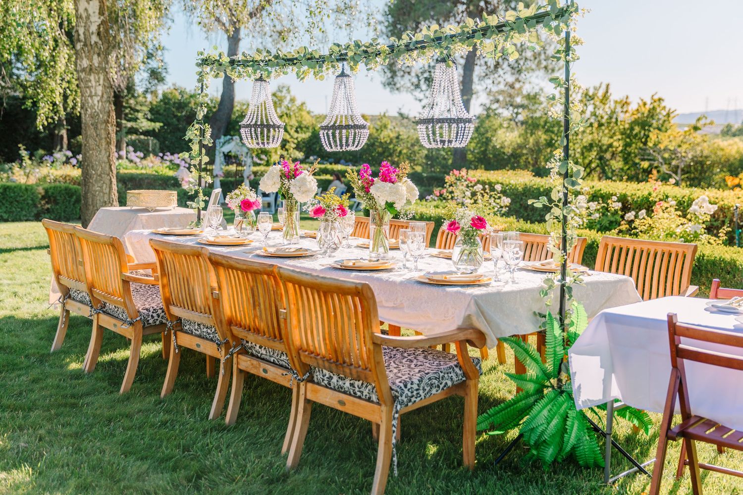 Beautiful wedding tablescape dining tables set up on a lawn with elegant place settings under a shaded tree canopy.