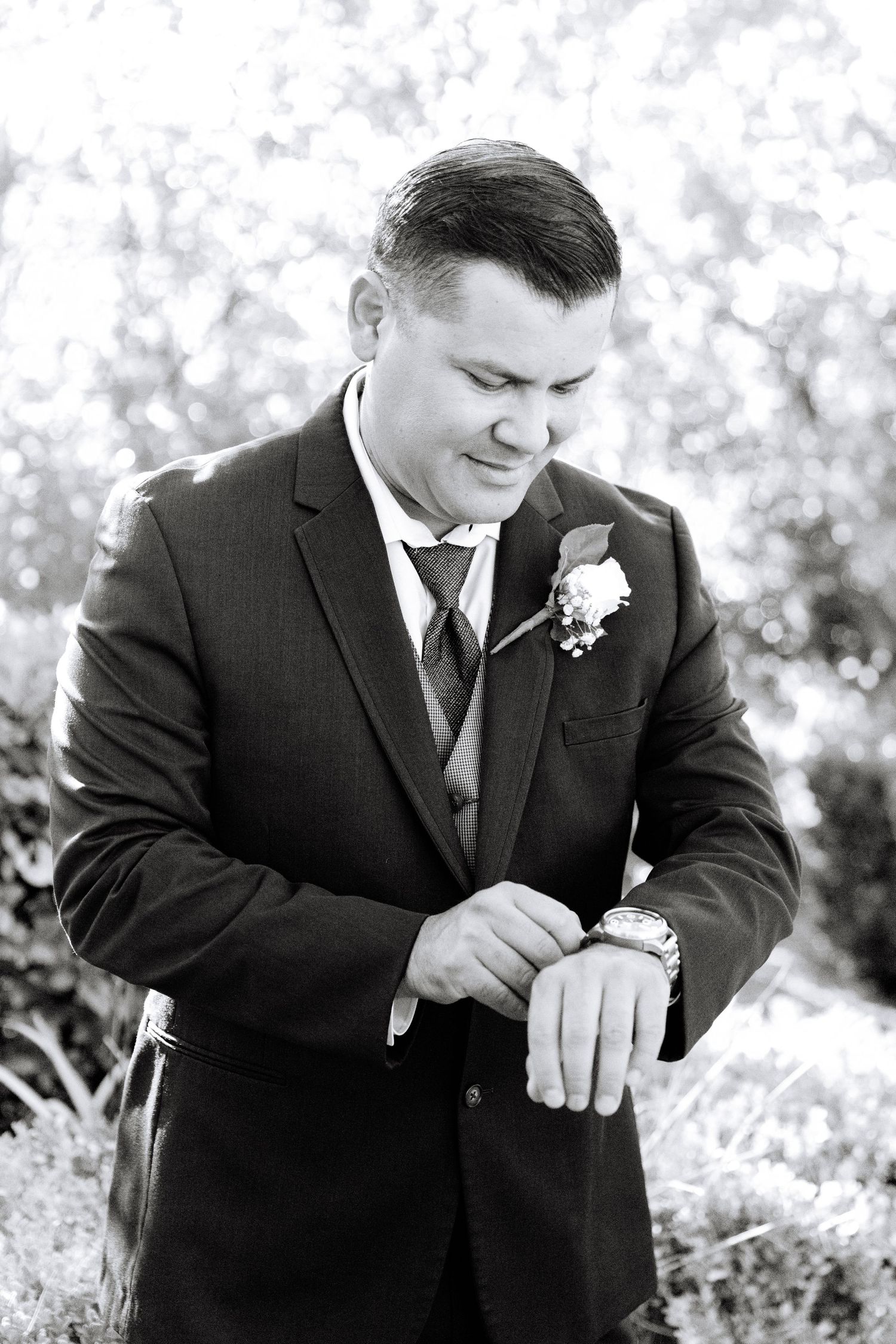 A groom in a black suit adjusts his sleeve while sitting outdoors in a natural setting.