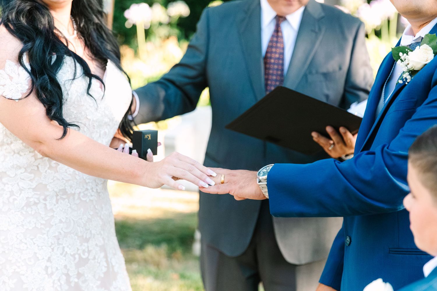 Couple holding hands during outdoor wedding ceremony while officiant holds black folder in natural setting.
