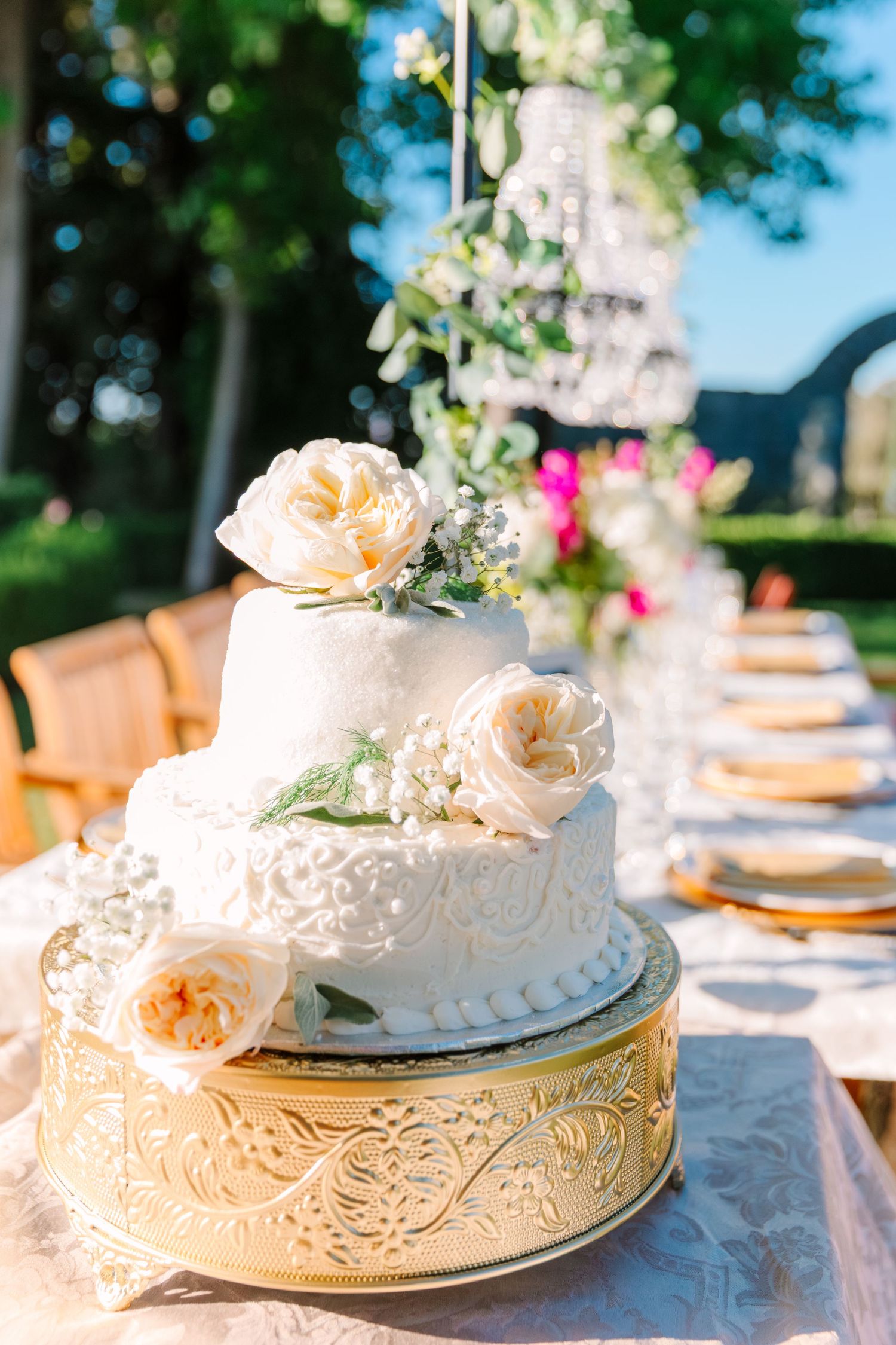 Elegant white wedding cakes adorned with lace details and floral decorations displayed on ornate plates at an outdoor reception.
