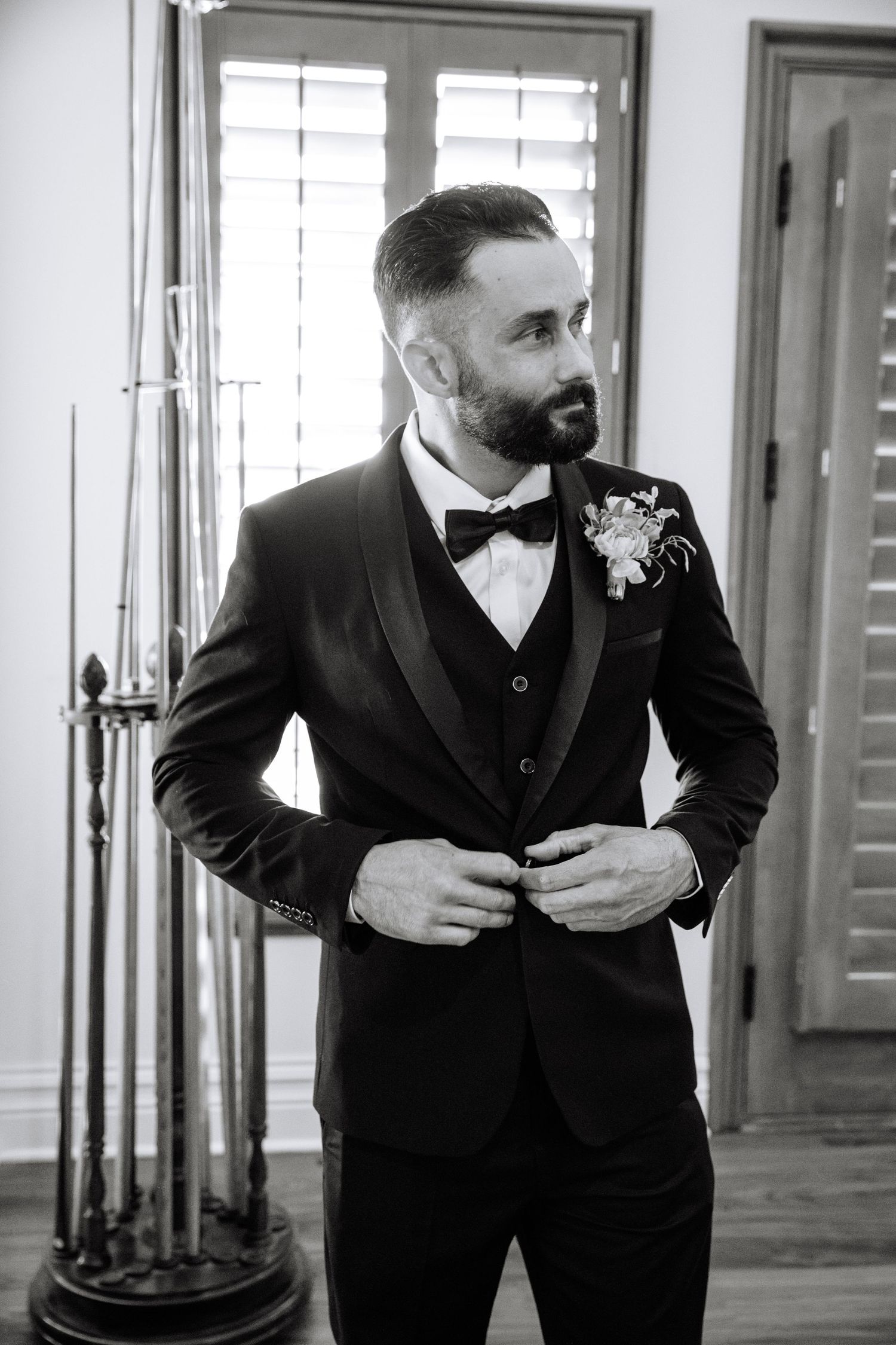 A groom in a classic black tuxedo and bow tie adjusts his jacket while getting ready in an elegant room with shuttered windows.