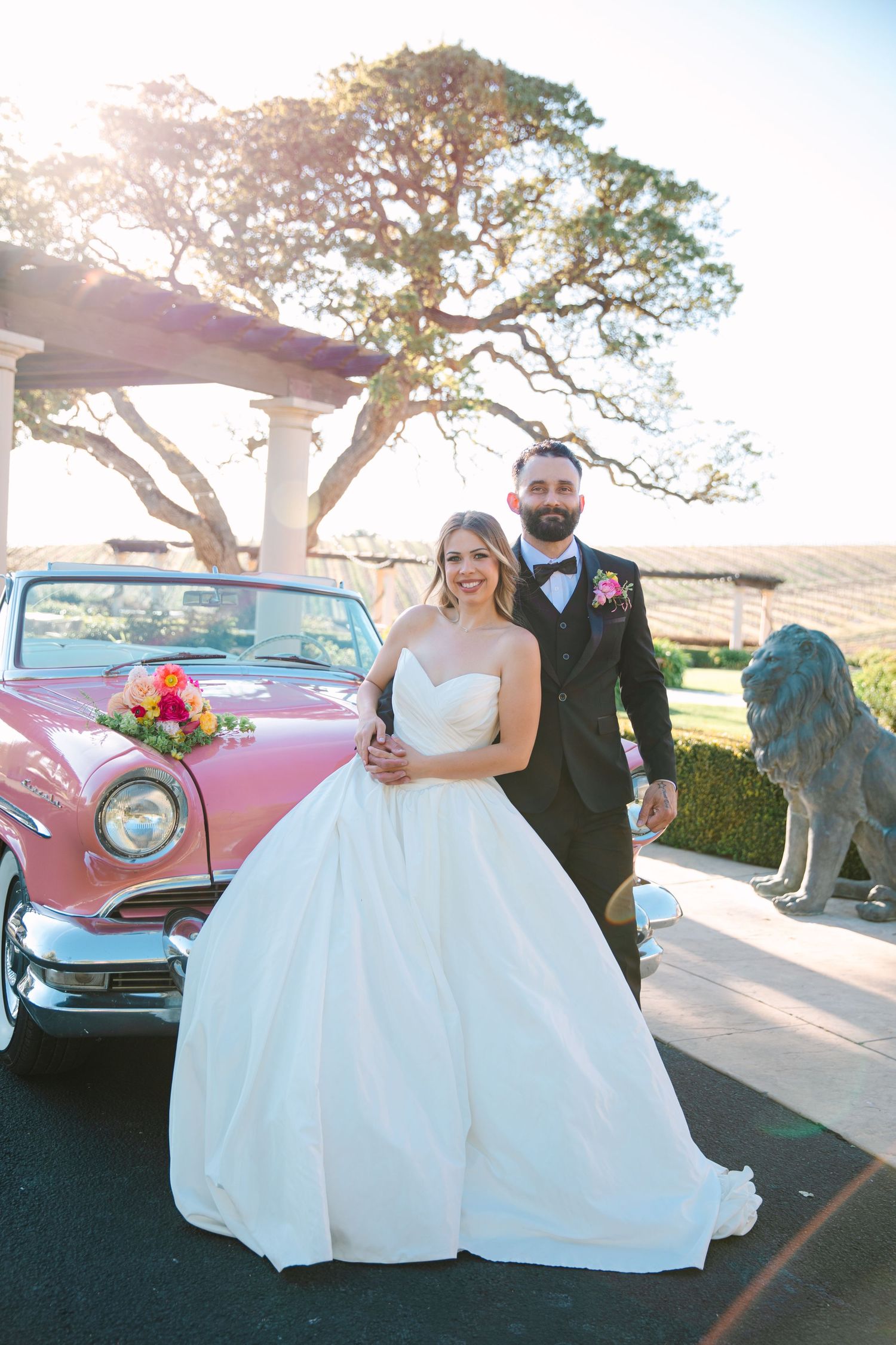 A classic pink vintage car parked at an elegant wedding venue with a stone lion statue.