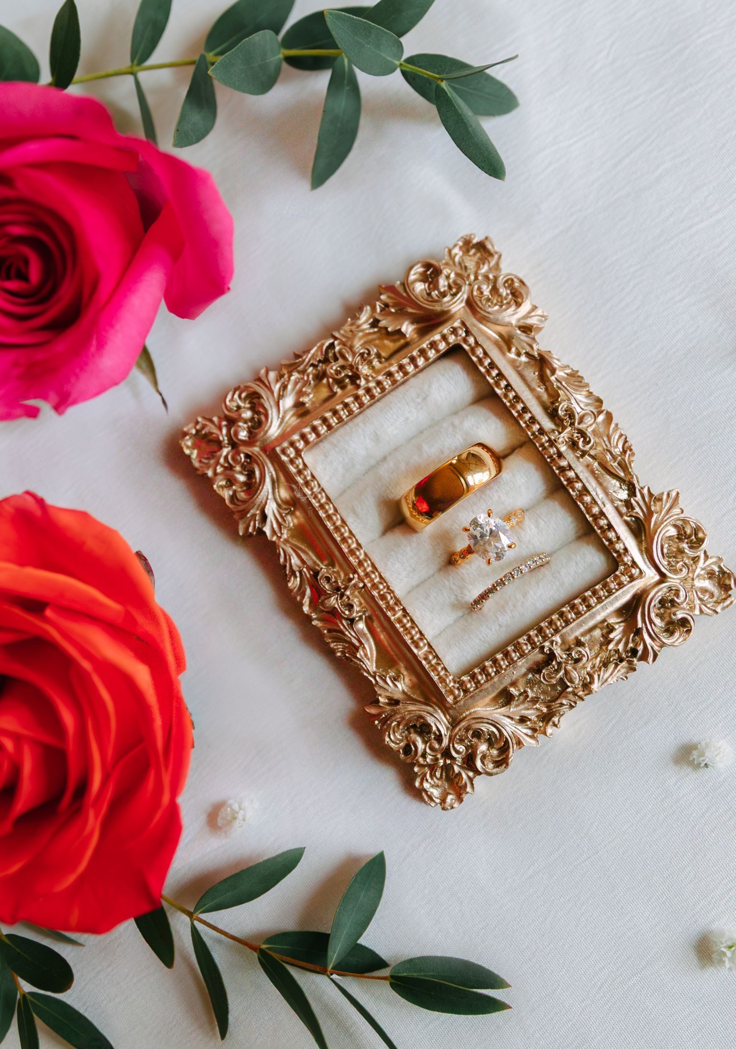 Wedding rings displayed in ornate gold vintage frame with red roses and eucalyptus leaves on white background.