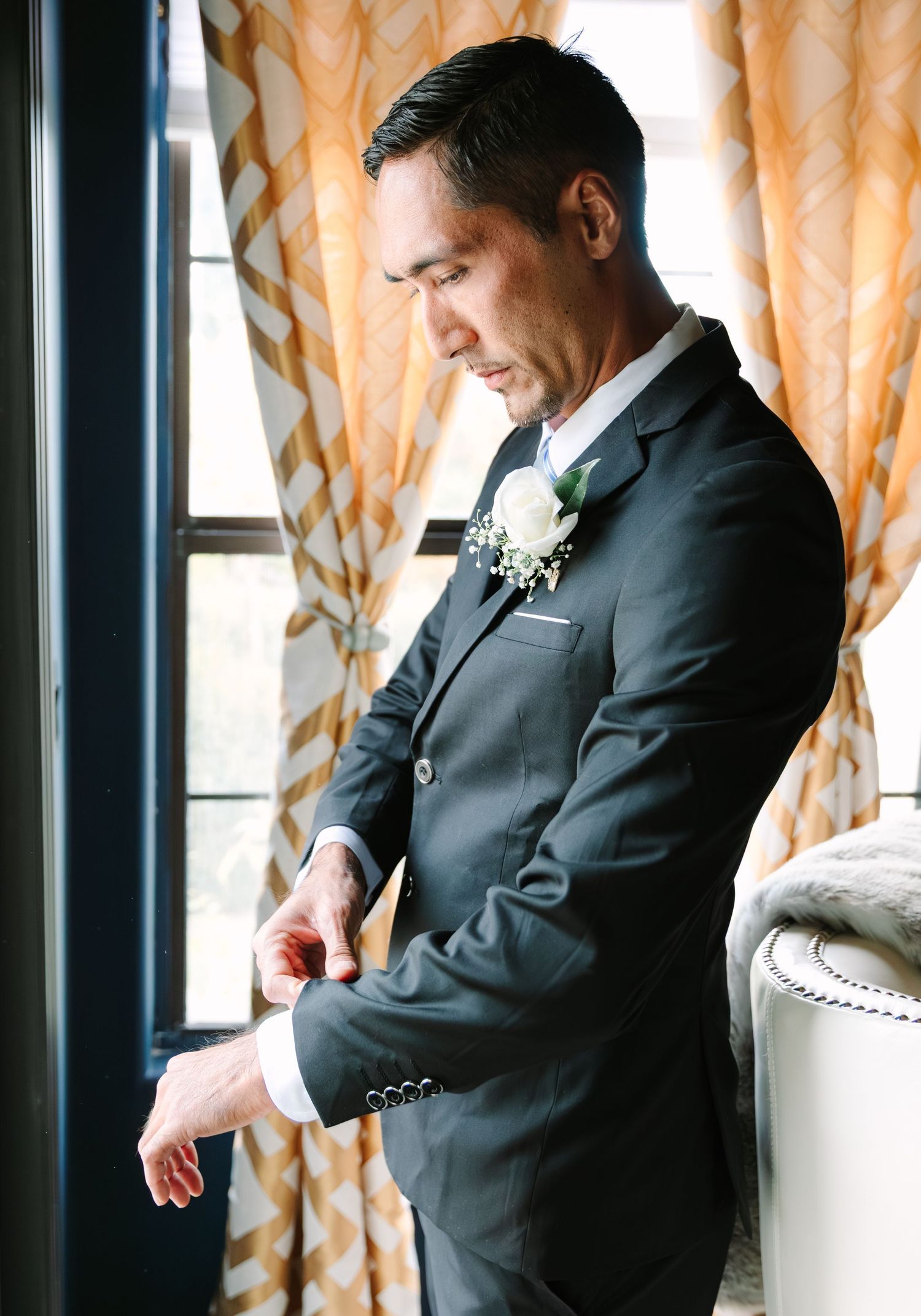 A groom in a classic black suit adjusts his tie while standing by a window with orange curtains.