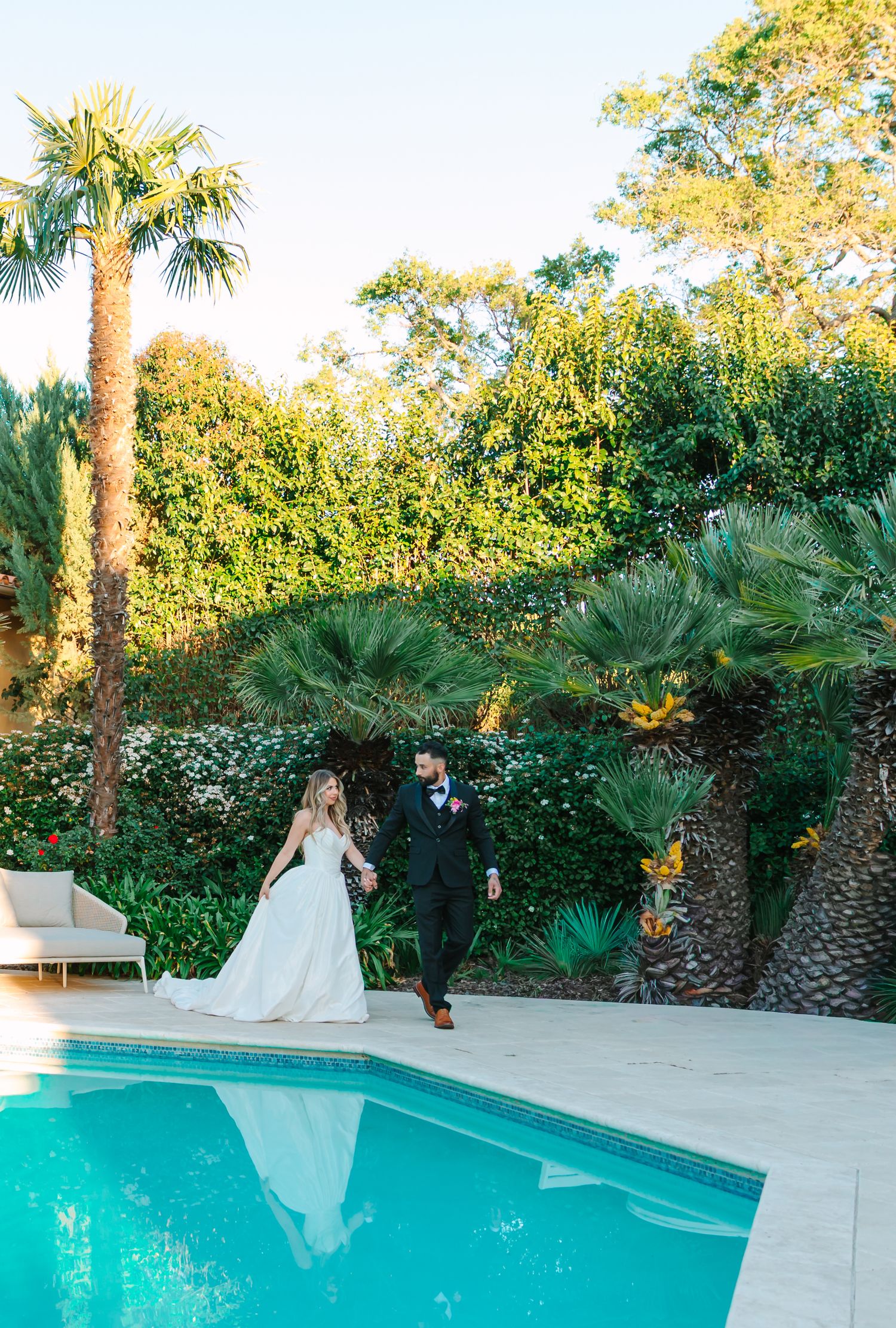 Couple in formal attire stands near a luxurious swimming pool surrounded by palm trees and lush greenery at sunset.