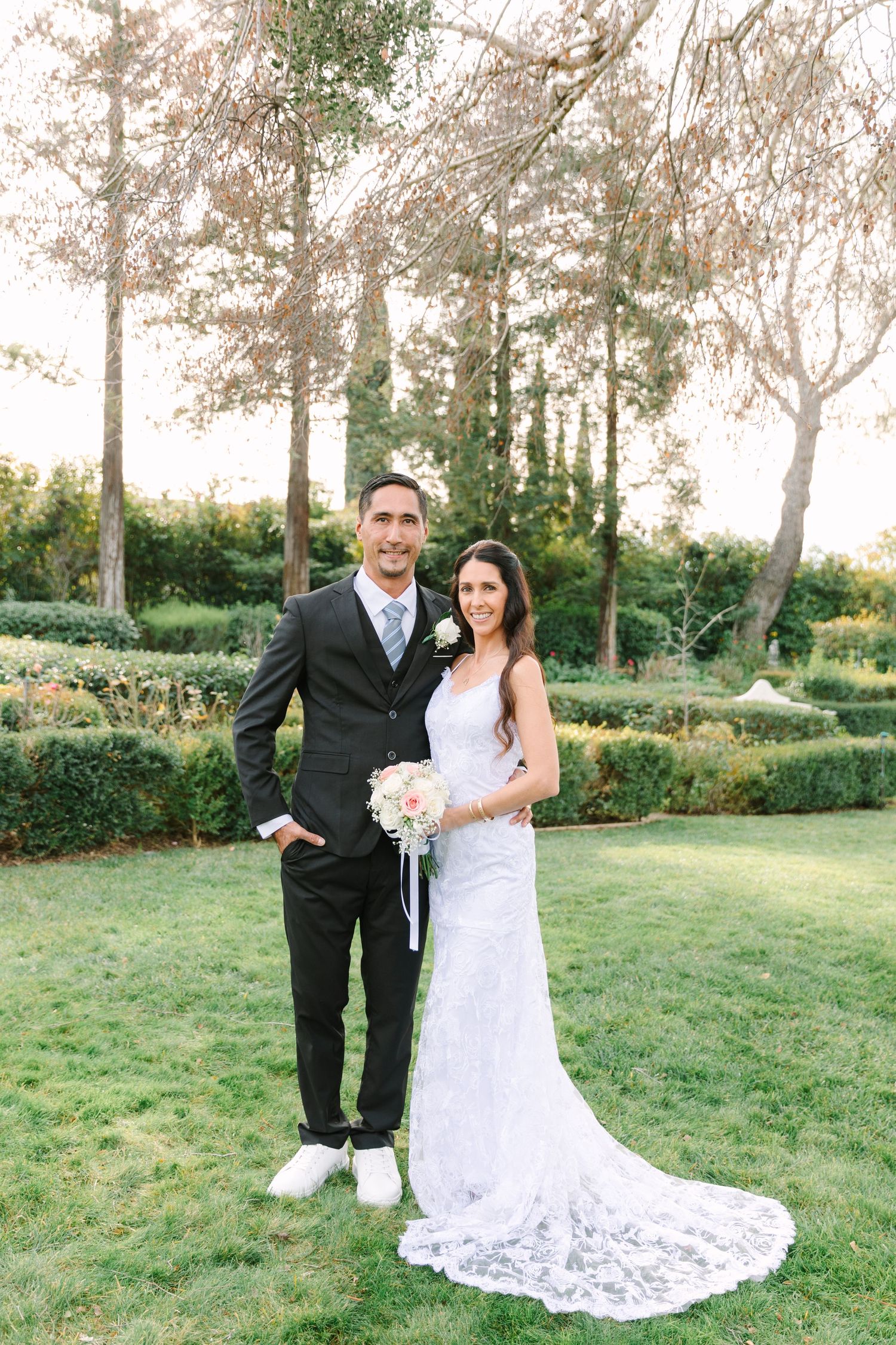 A couple poses in formal wedding attire on a grassy lawn with trees in the background.