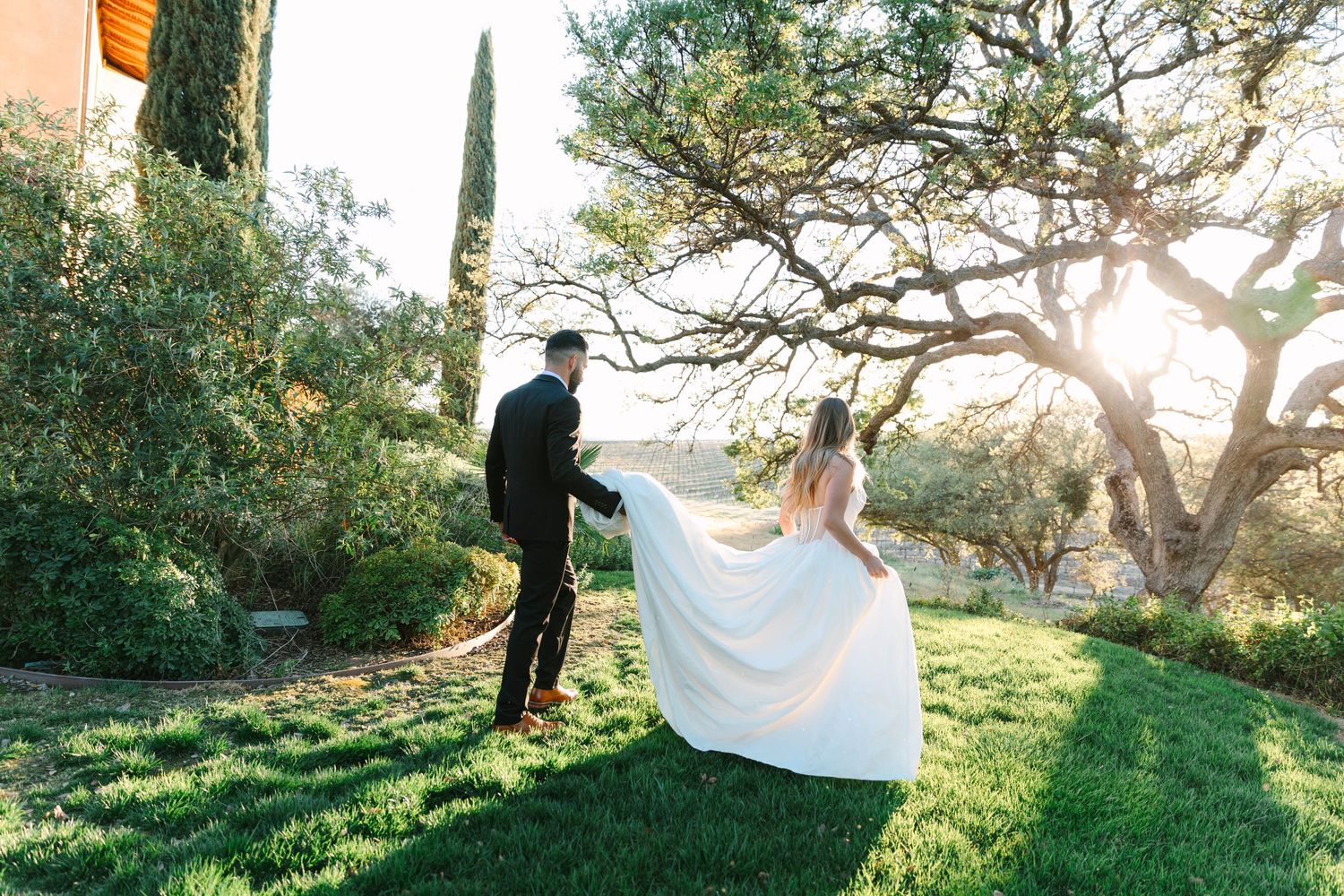 Couple in wedding attire walks through sunlit garden path with trees casting dappled shadows at sunset.