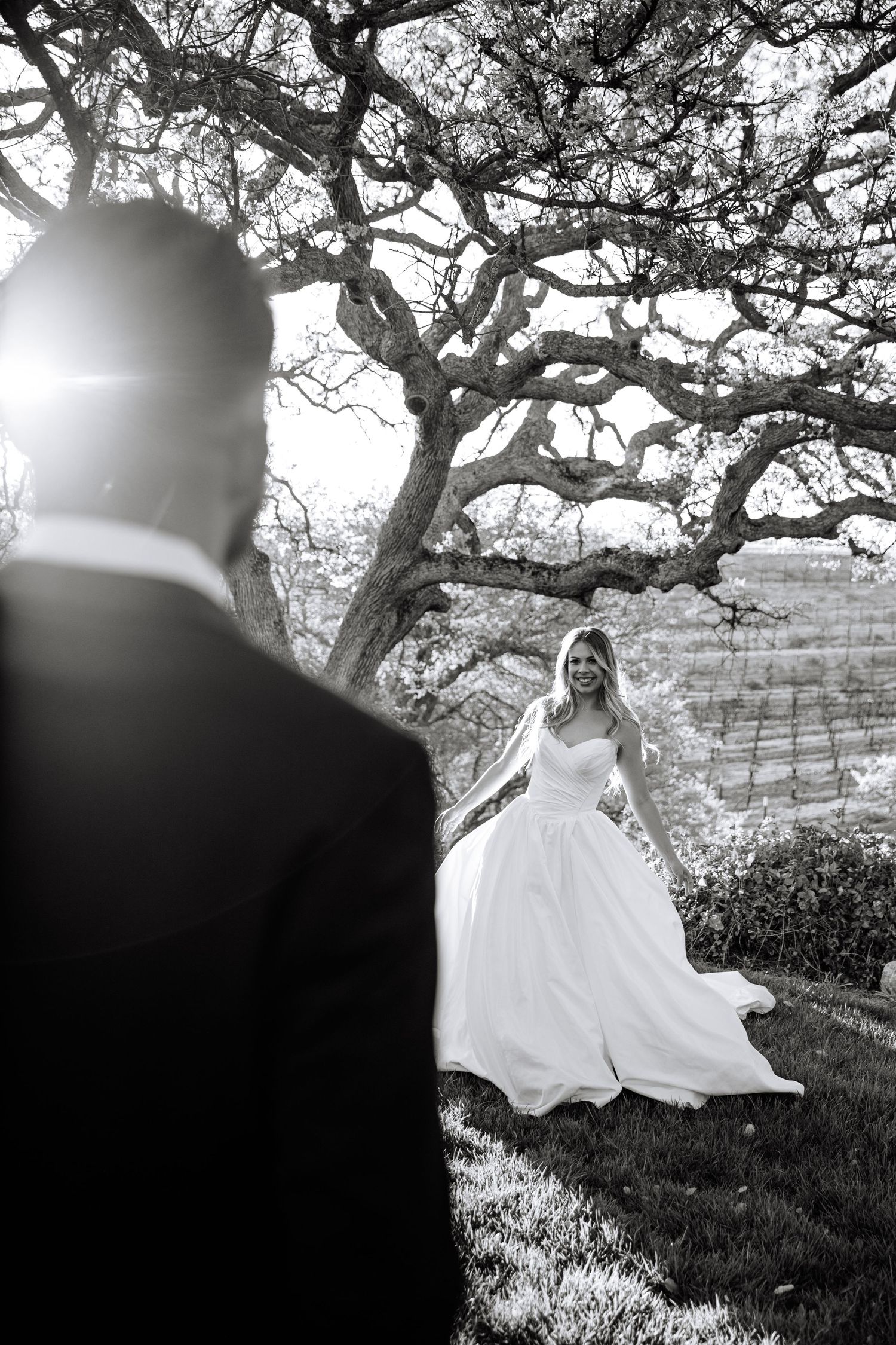 A bride in a flowing white wedding dress twirls joyfully under a large tree branch in a black and white photograph.
