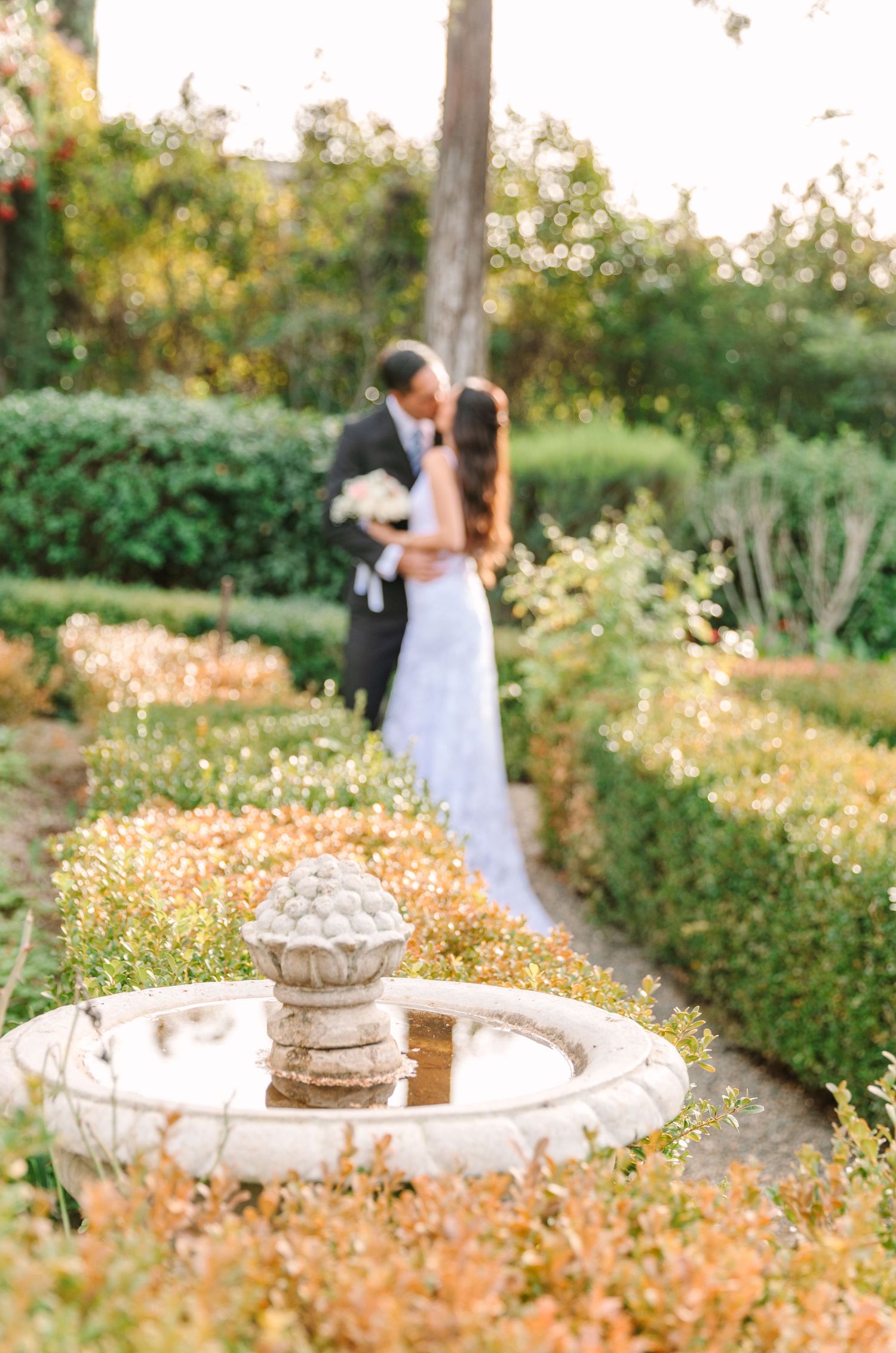 A couple embraces in a romantic garden setting with stone planters and wildflowers during golden hour.