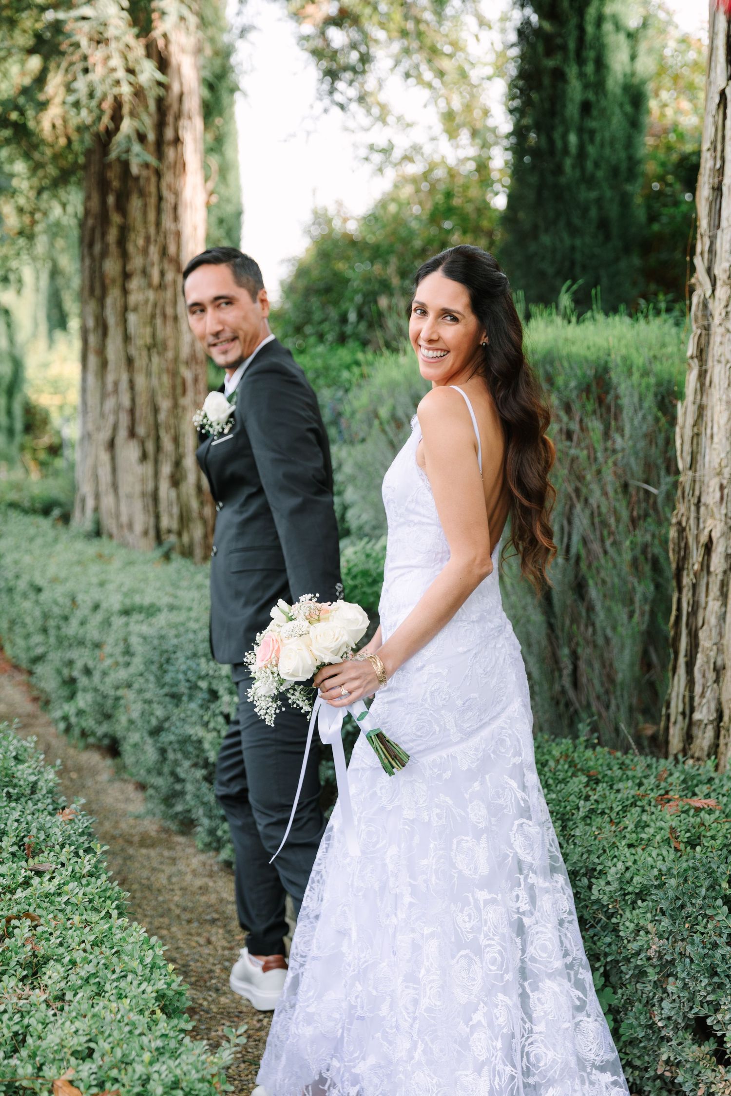 A romantic wedding photo of a couple posing in a garden path lined with cypress trees and greenery.