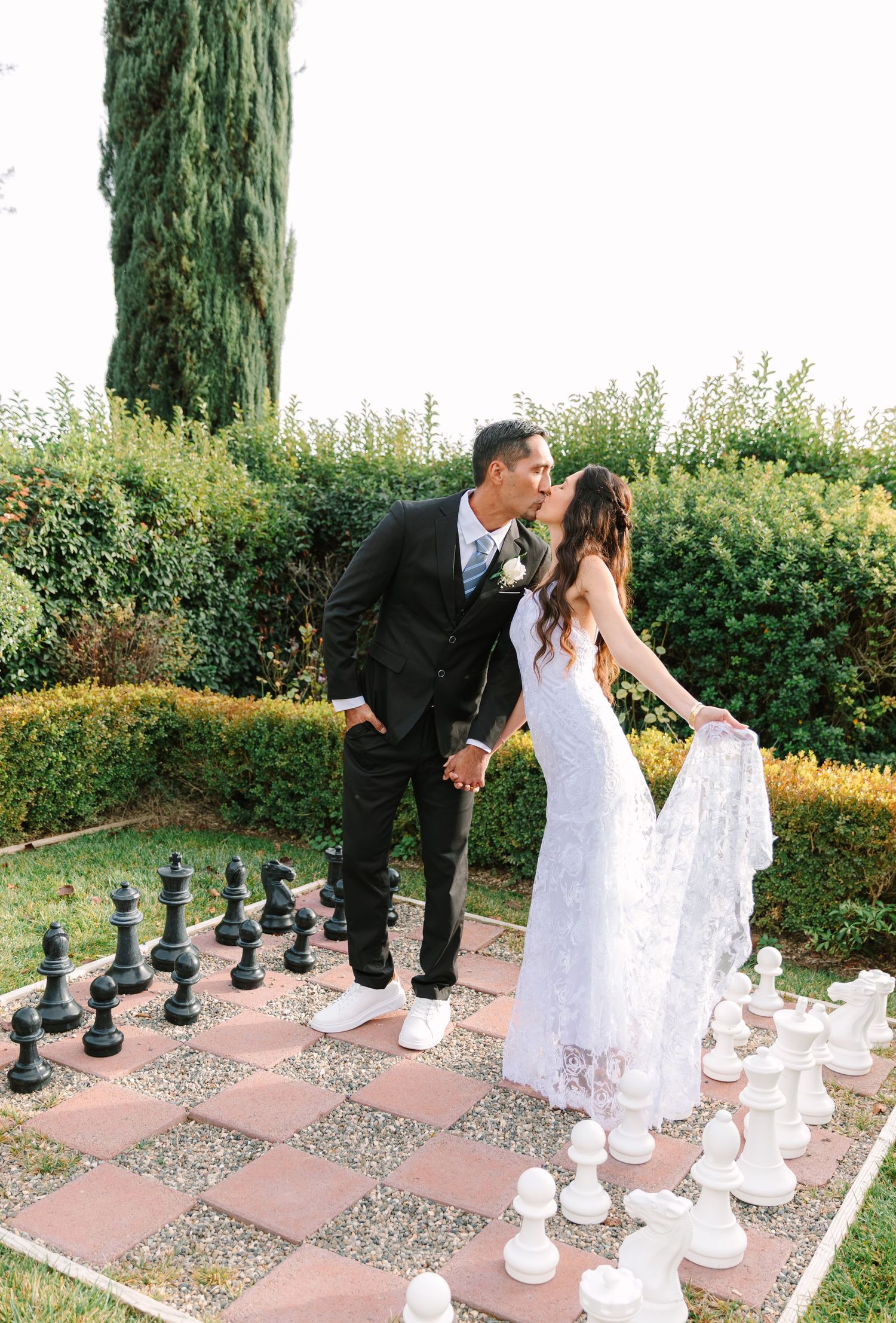 A couple in wedding attire plays a game of life-sized chess on an outdoor patio surrounded by greenery.