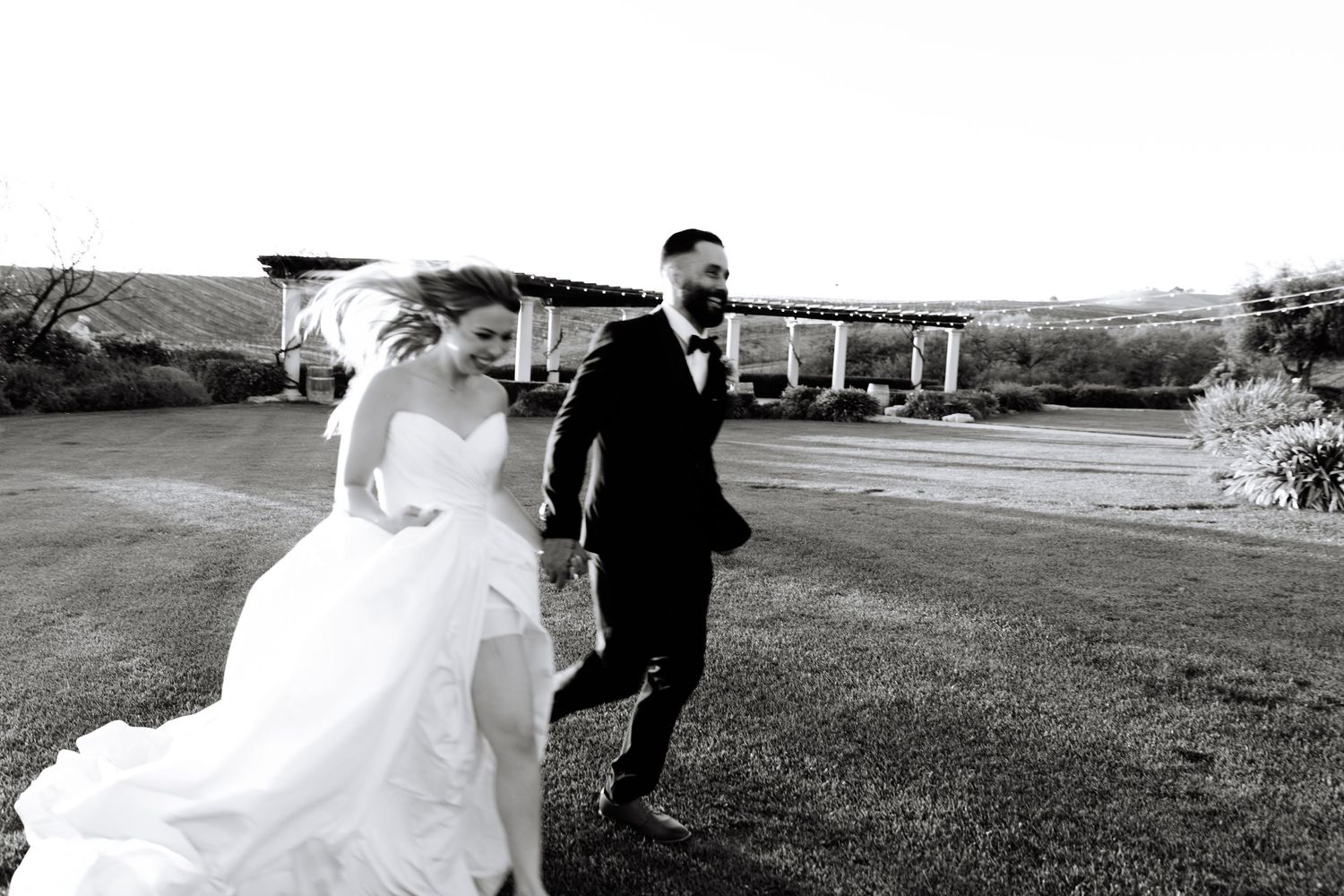 A couple in wedding attire walks through a grassy field at sunset with the bride's dress flowing in the wind.