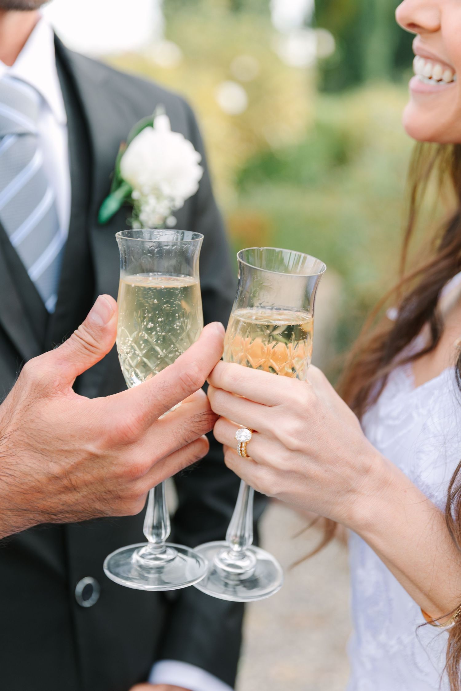 A couple toasts with champagne flutes at a wedding celebration, showing hands with rings and formal attire.