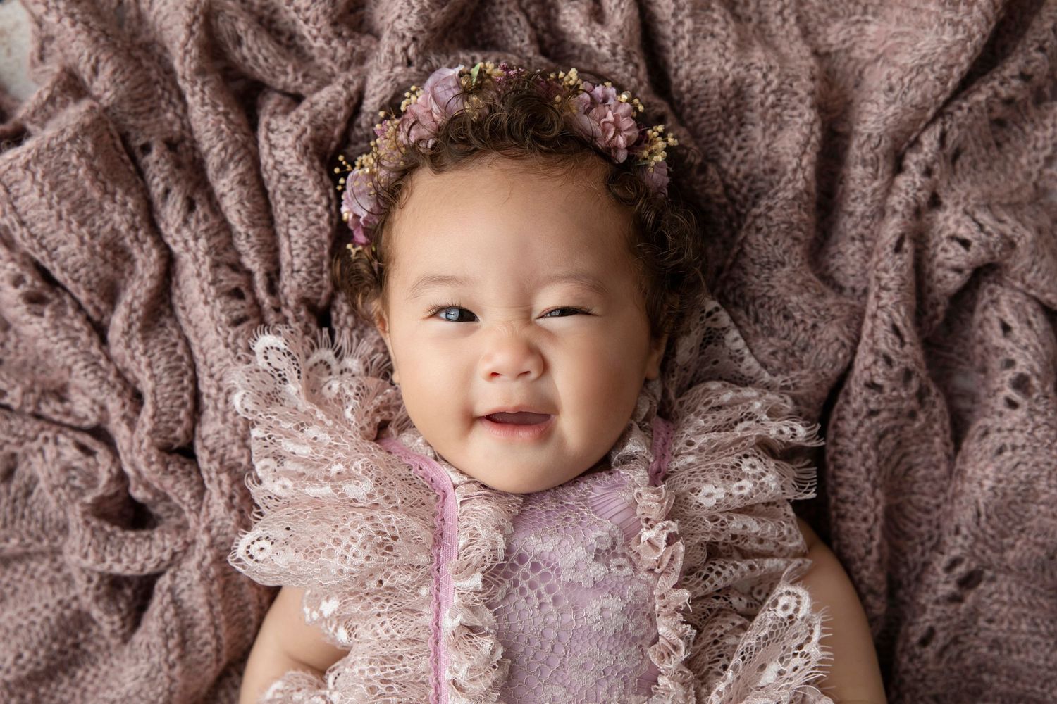 Happy baby wearing lace dress lying on textured mauve blanket with floral crown.
