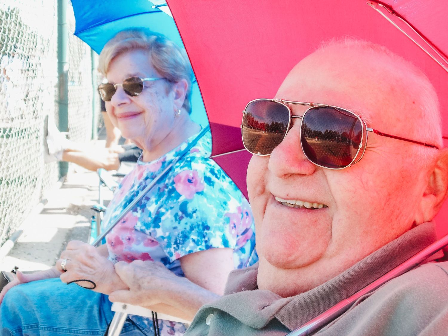 Two senior individuals wearing sunglasses sit together under a bright red umbrella on a sunny day.