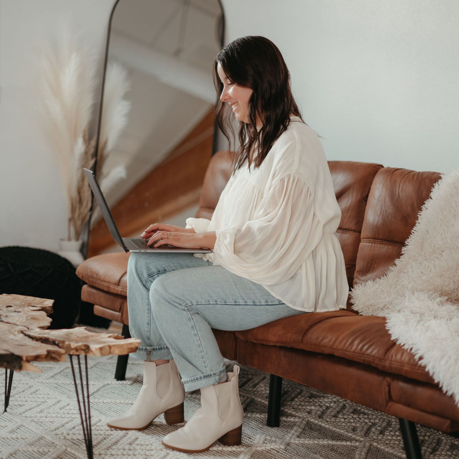 Person in white blouse and jeans sits on cognac leather couch working on laptop with coffee table nearby.