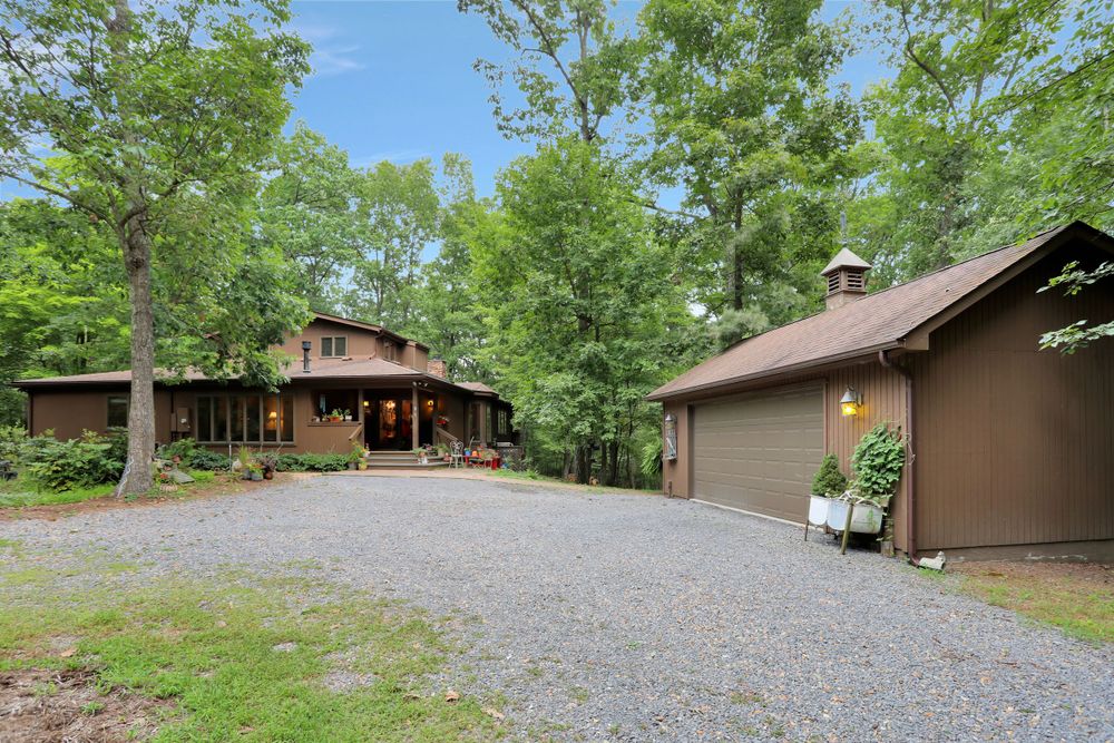 Rustic brown cabin nestled in wooded setting with gravel driveway and detached garage.