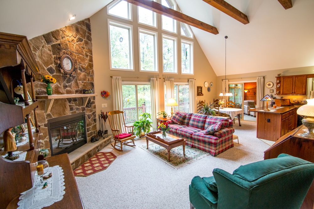 Spacious cabin interior featuring stone fireplace, high windows and plaid furniture in an open floor plan.