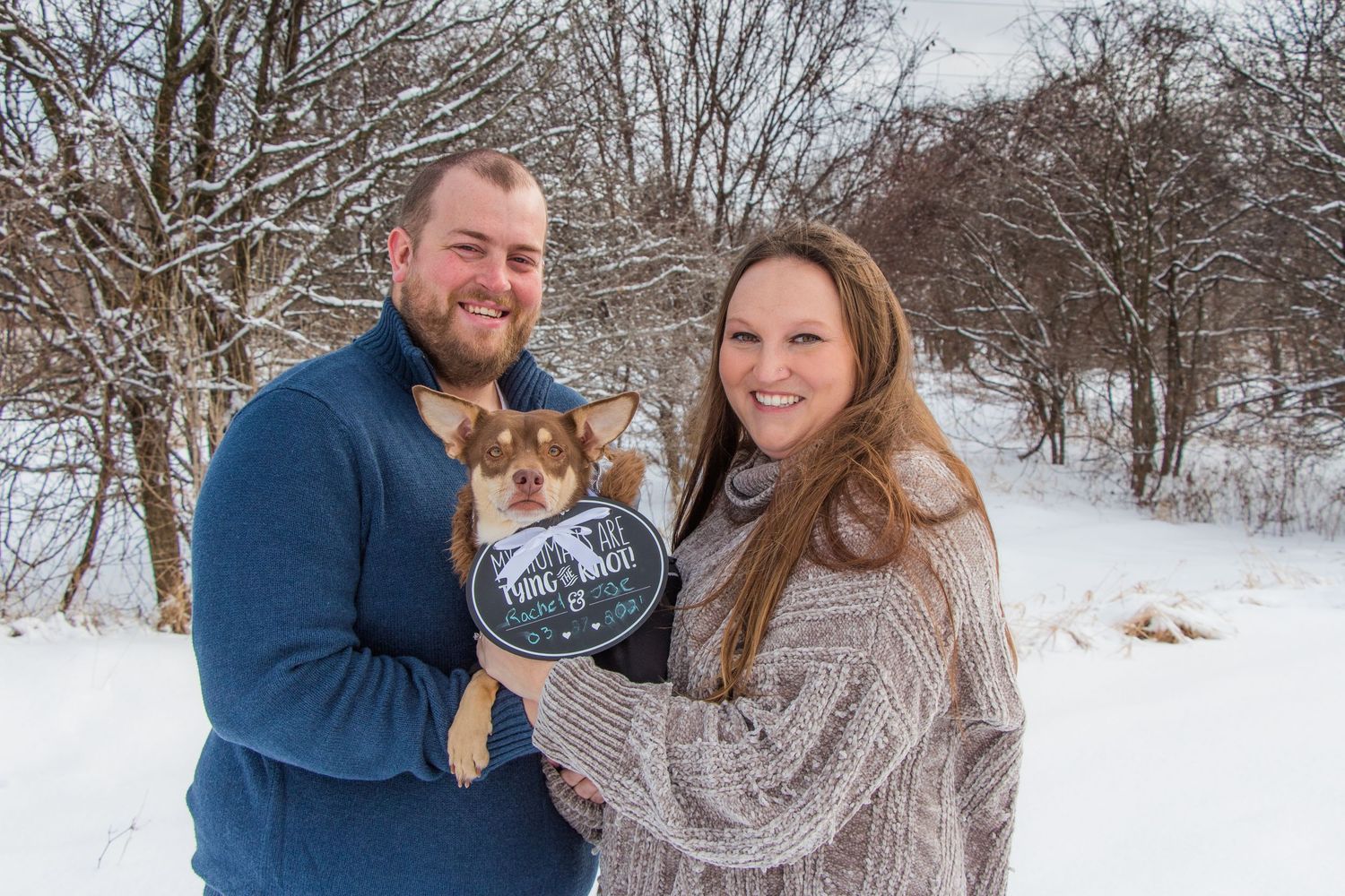 A couple in winter clothing holding their pet dog against a snowy landscape background.