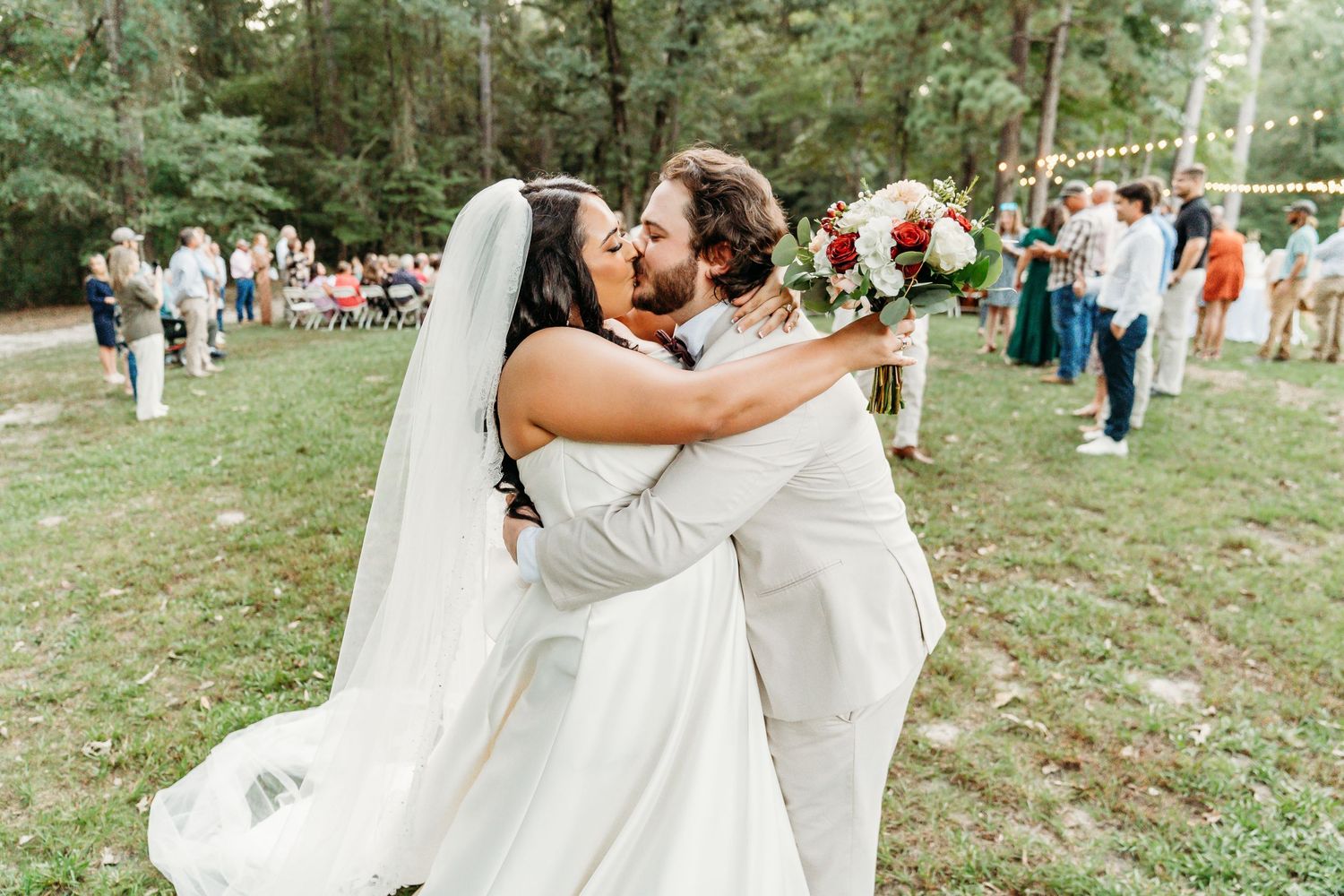 A couple shares a romantic kiss at an outdoor wedding ceremony while guests look on in a wooded setting.