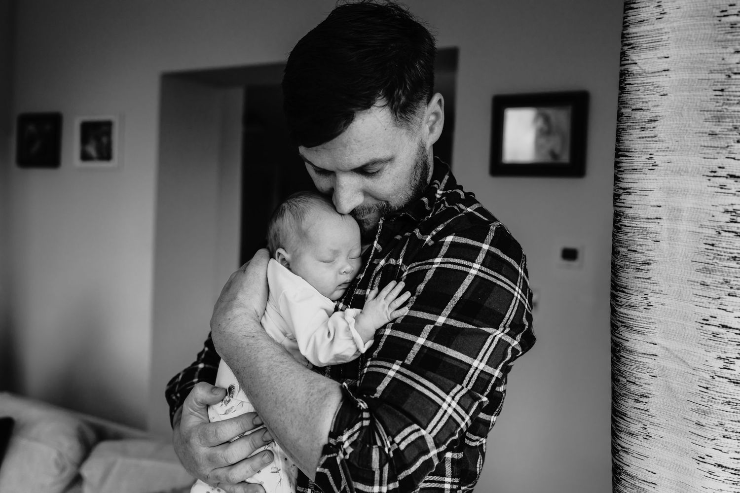 Person wearing a plaid shirt holds and kisses a baby in a tender black and white indoor photograph.