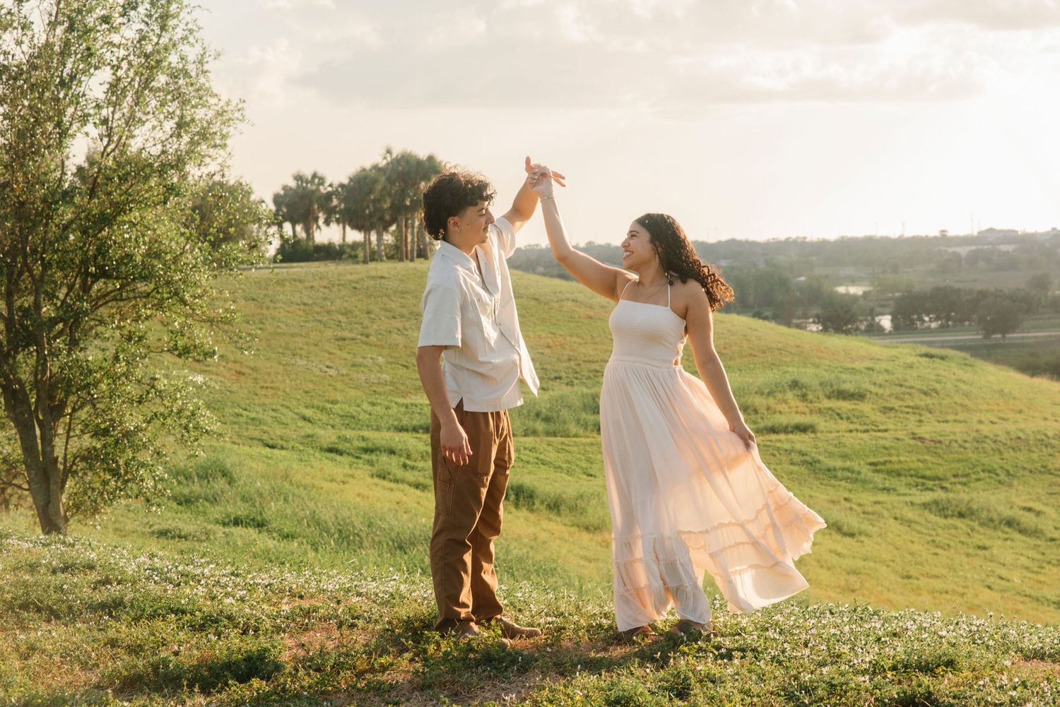A couple dances together on a hilltop meadow during a romantic sunset photoshoot.