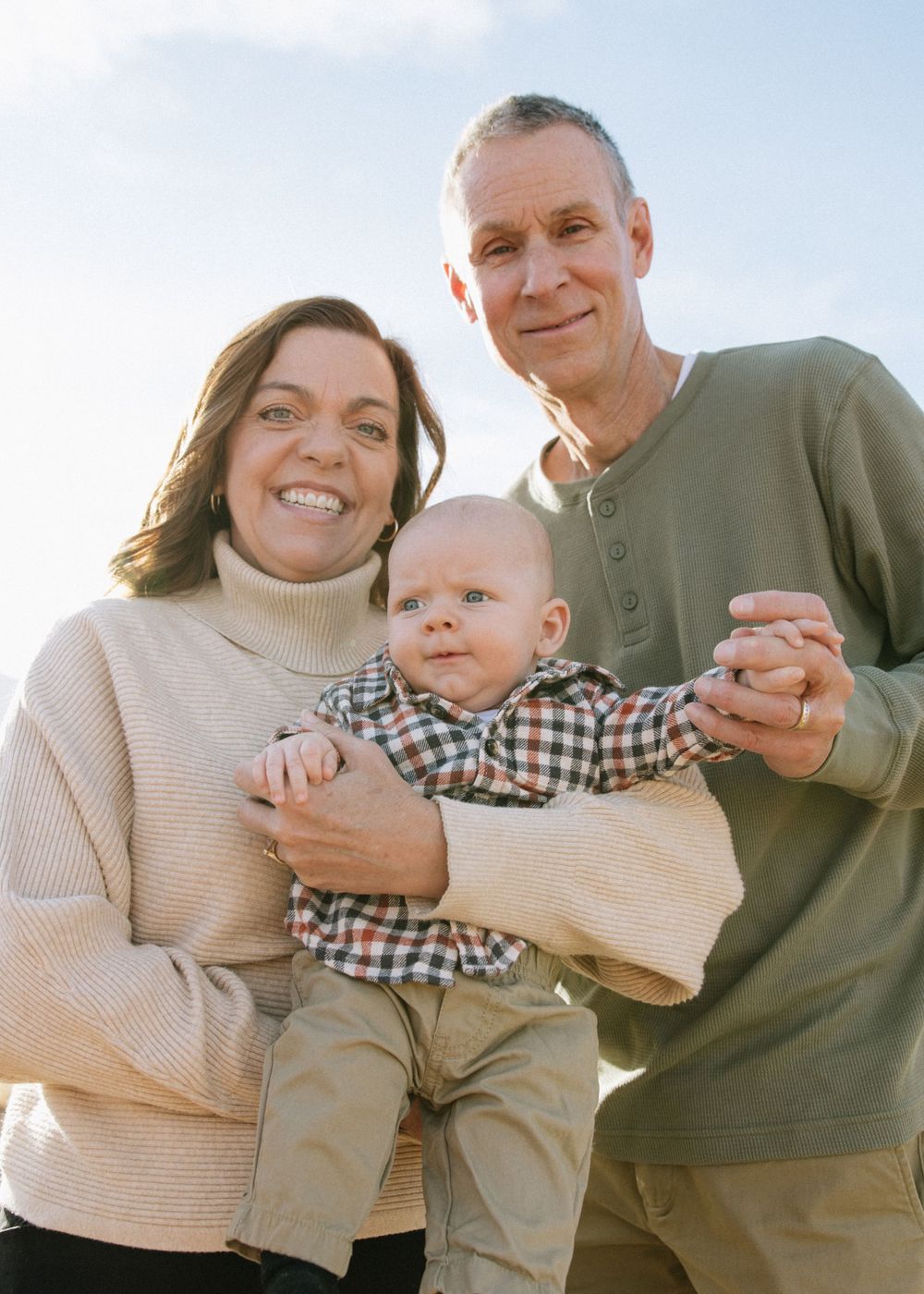 A family portrait outdoors with a baby wearing a plaid shirt on a sunny day.