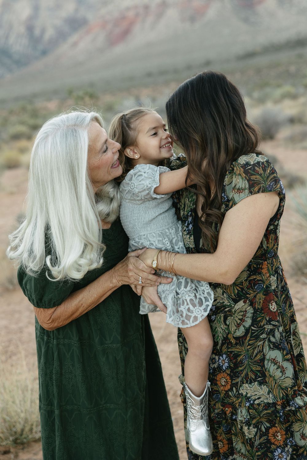 A loving family embraces outdoors against a desert mountain backdrop in matching dark green and floral outfits.