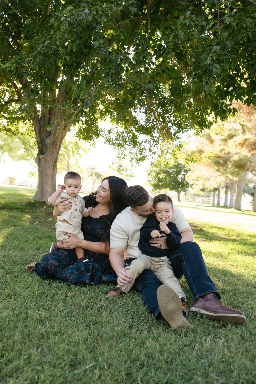 Family members playfully pile on top of each other while laying in green grass under shady trees on a sunny day.