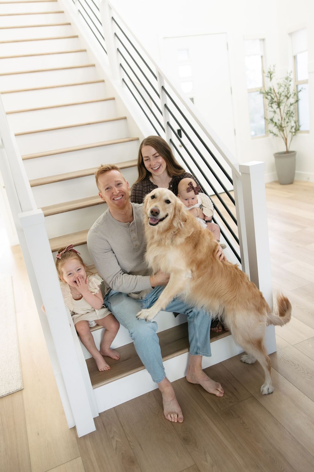 A family sits together on white wooden stairs with their friendly golden retriever in a bright modern home interior.