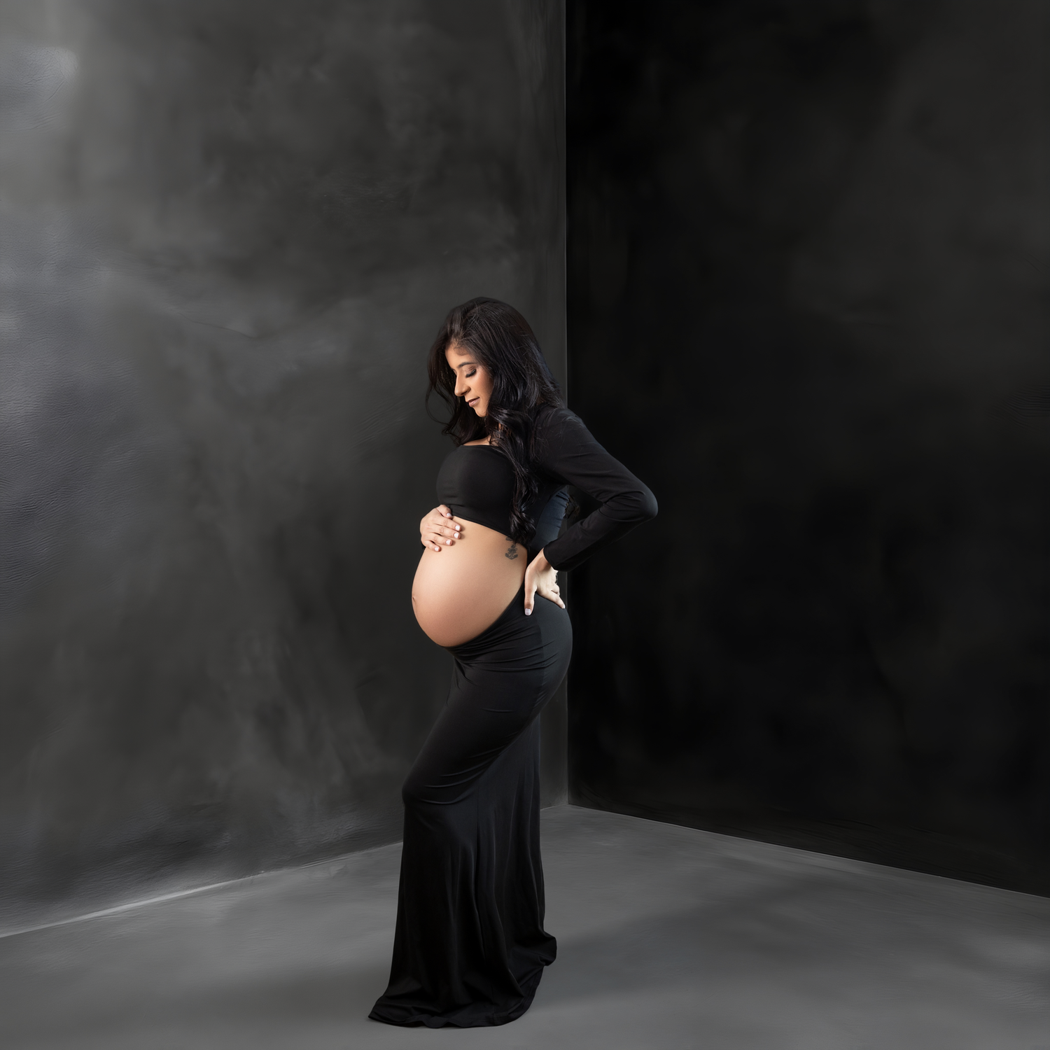 A pregnant figure in a cream-colored flowing dress poses gracefully against a textured mauve backdrop.