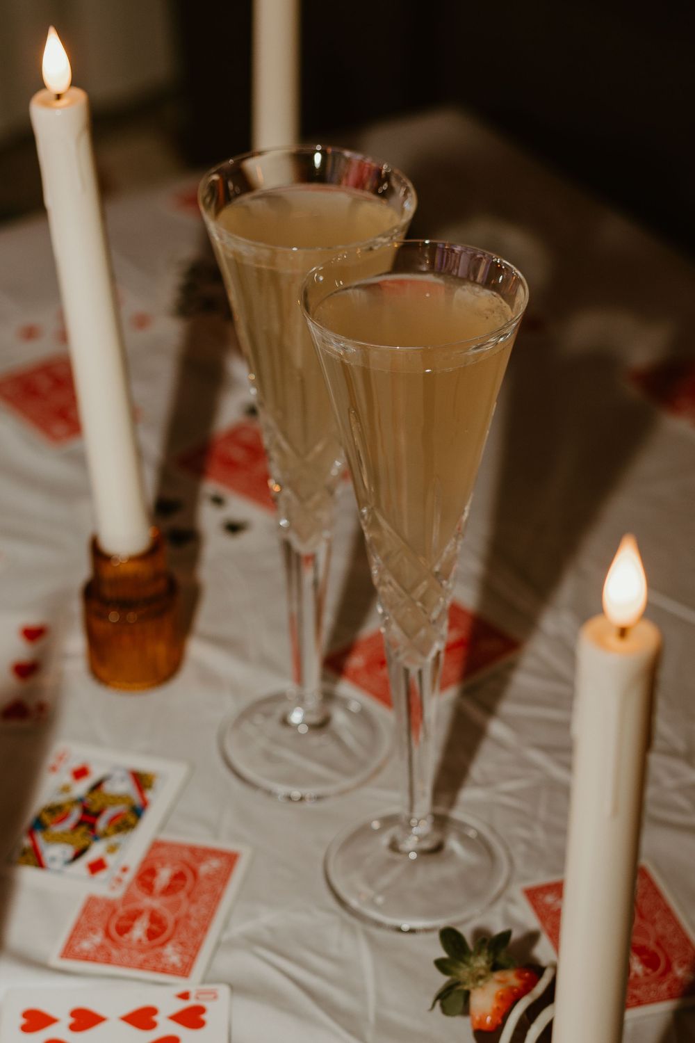Two champagne glasses and playing cards set on a white tablecloth with lit candles creating a romantic atmosphere.
