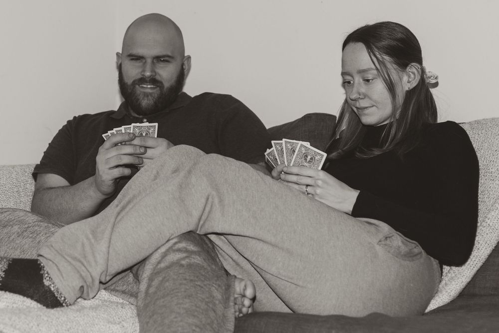 A couple relaxes on a sofa while playing cards together in black and white.
