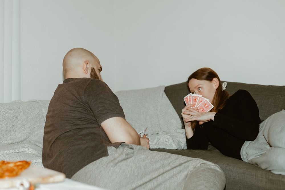 A couple lounges on a grey couch playing cards together in a cozy indoor setting.
