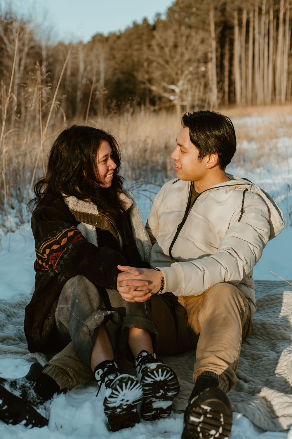 Couple sits together on snowy ground during winter forest photoshoot.