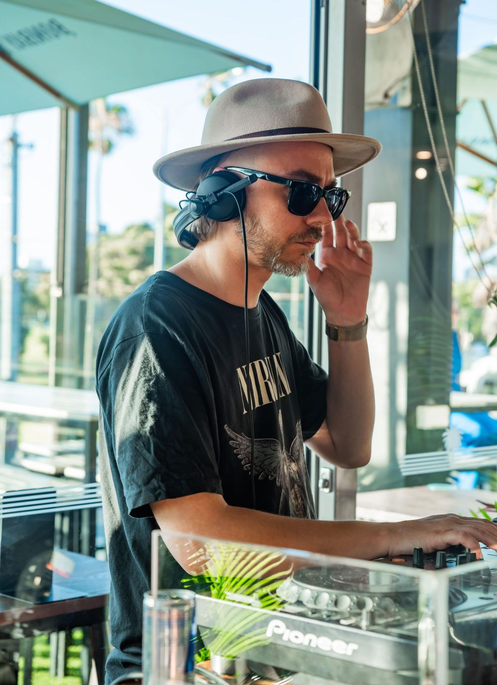 DJ performing at outdoor venue wearing fedora and sunglasses.