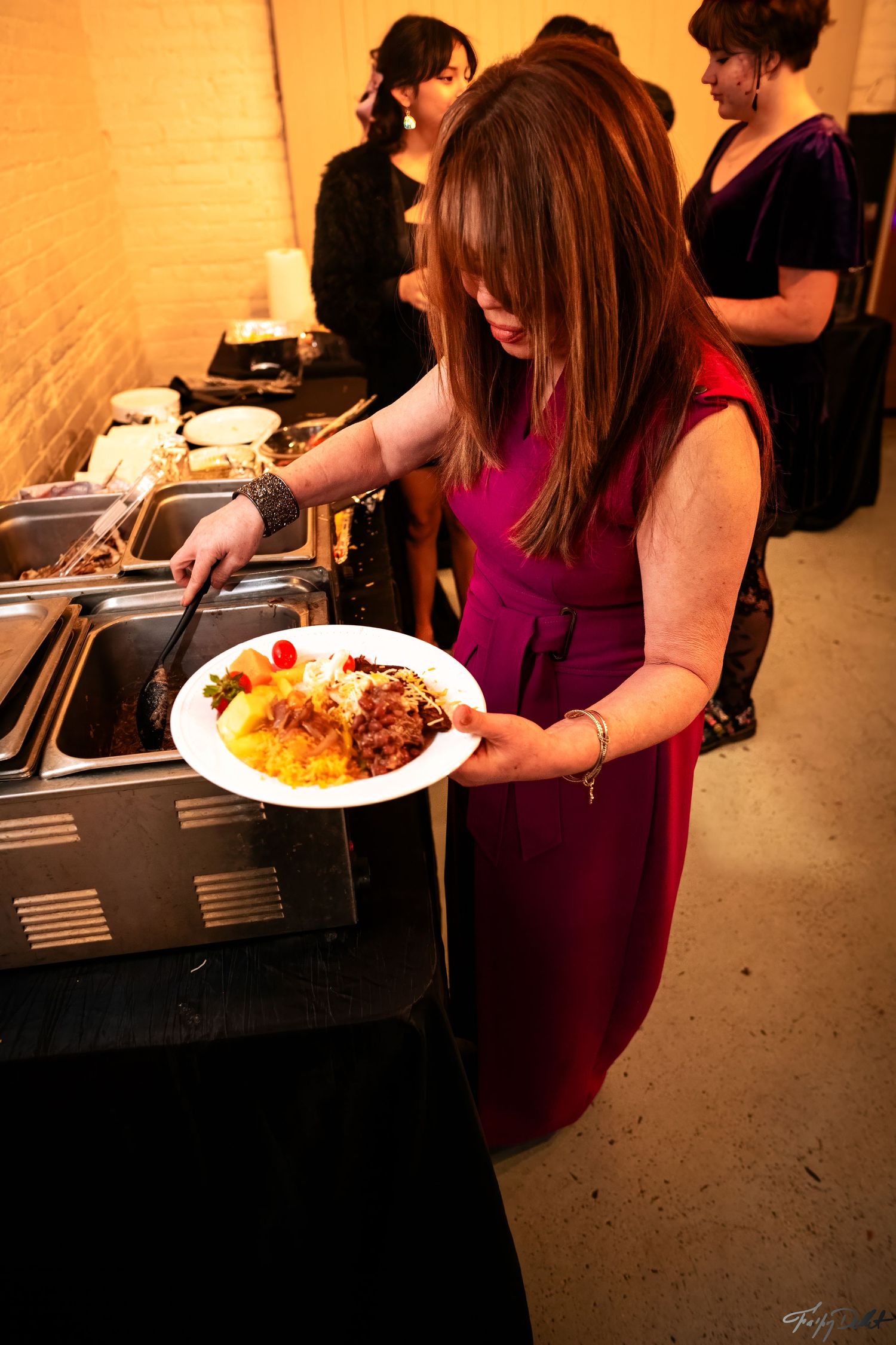 A guest in a burgundy dress serves food from a buffet line at an event.