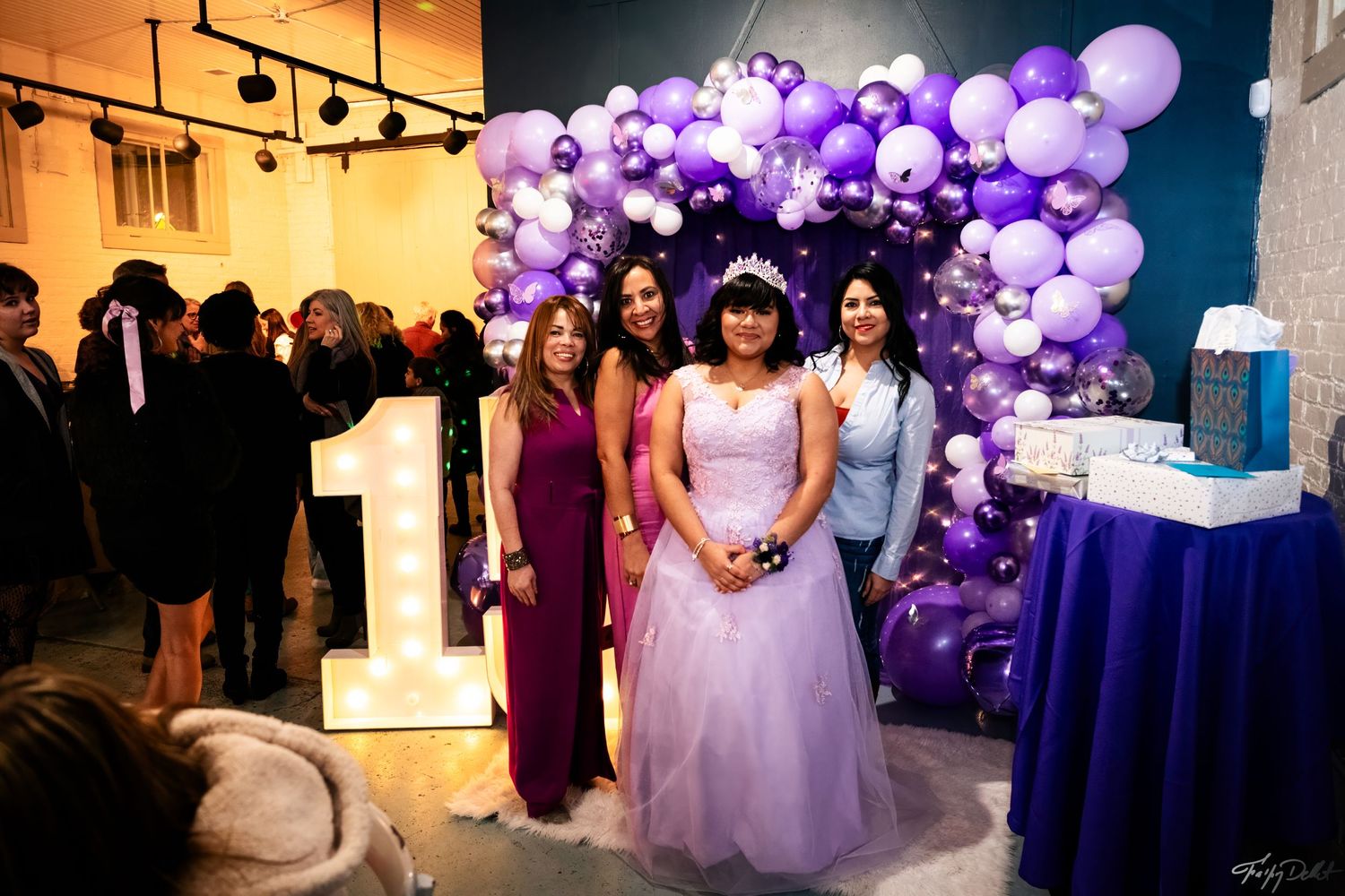 A celebration setup with purple balloon arch, illuminated number display, and formal attire.