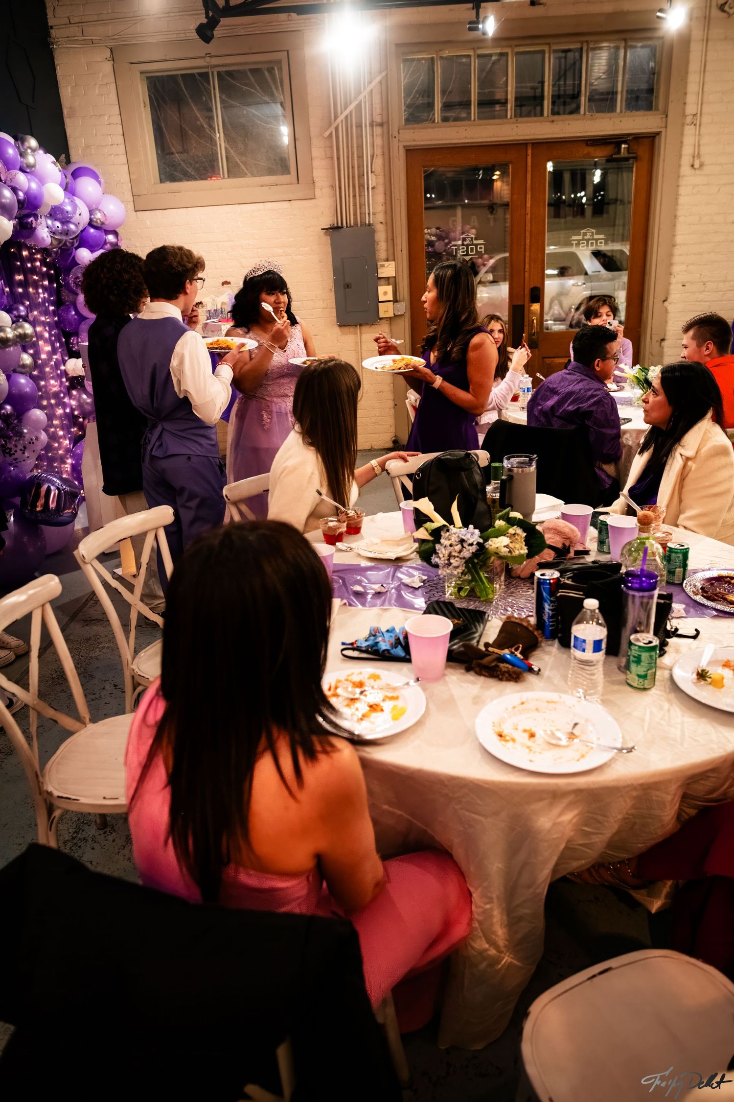 A group gathers around decorated tables at a celebration with purple balloons and decor.