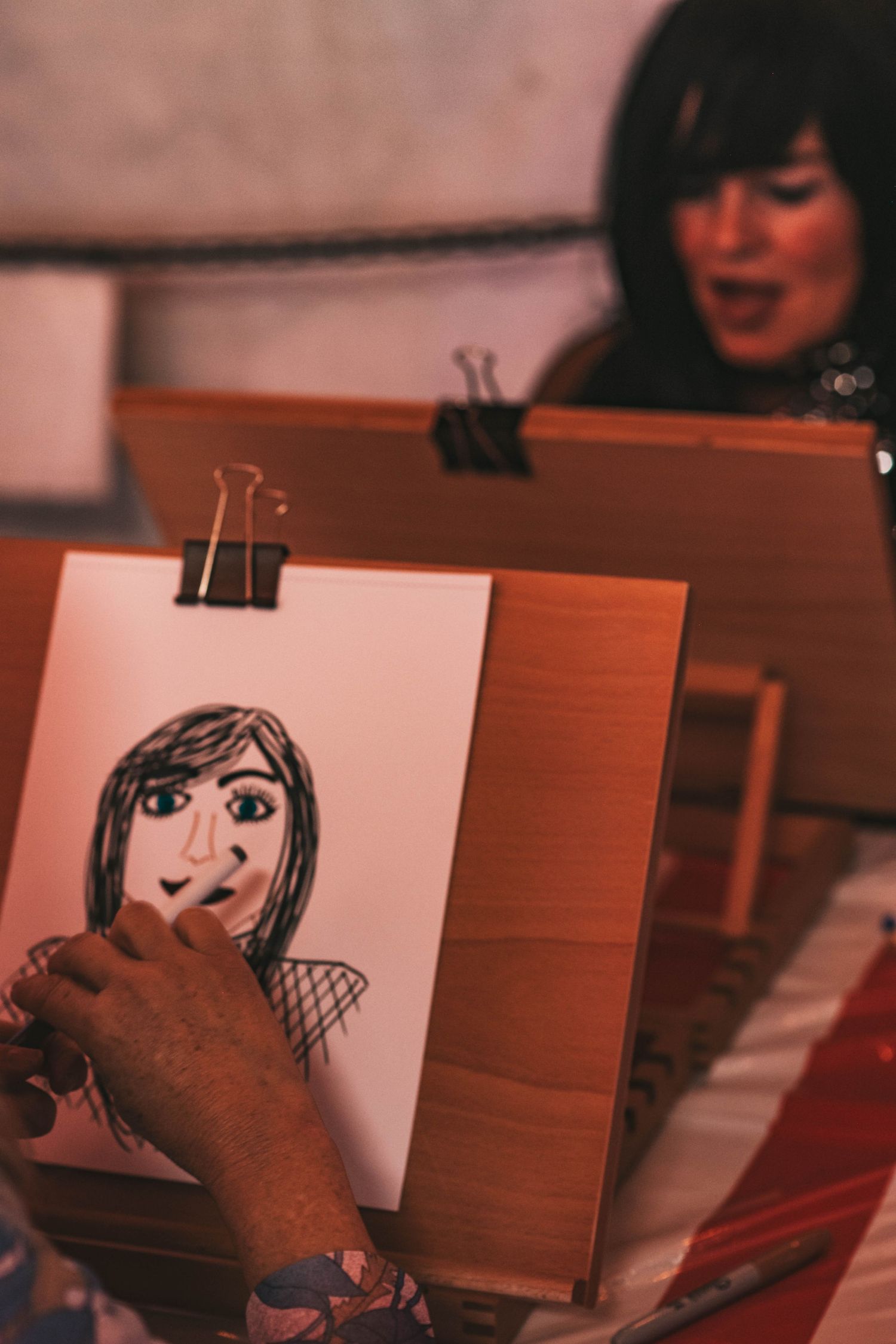 An artist sketches a simple portrait on paper held by binder clips on a wooden surface in warm lighting.